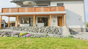 Una casa de dos pisos con un porche cubierto, un muro de contención de piedra y un pequeño jardín con flores.
