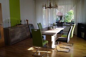 Uma área de refeições com mesa de madeira, cadeiras verdes, um lustre e vista para uma casa através de cortinas finas.