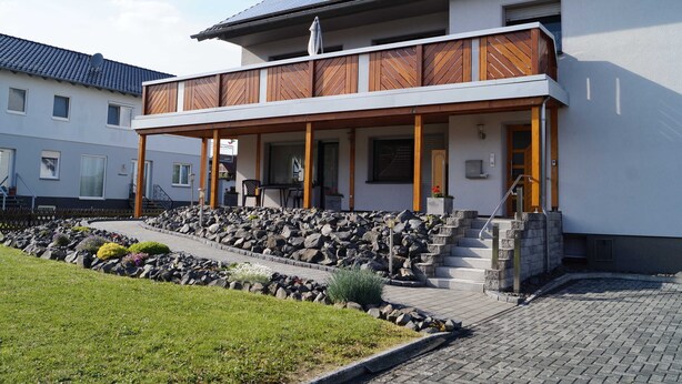 Una casa moderna de dos pisos con balcón, escalones de piedra y jardín con rocas y plantas.
