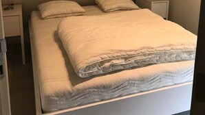 A small, single-bed room with a sloped ceiling, white walls, and a simple bedside table with a lamp. - Seabreeze 303 (Nieuwpoort)