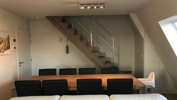 A modern dining area with a wooden table and black chairs, a staircase, and a white couch. - Seabreeze 303 (Nieuwpoort)