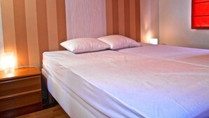 A neatly made bed with white linens and pillows, a wooden nightstand with a lamp, and striped wallpaper. - Marco Polo 0102 (Nieuwpoort)
