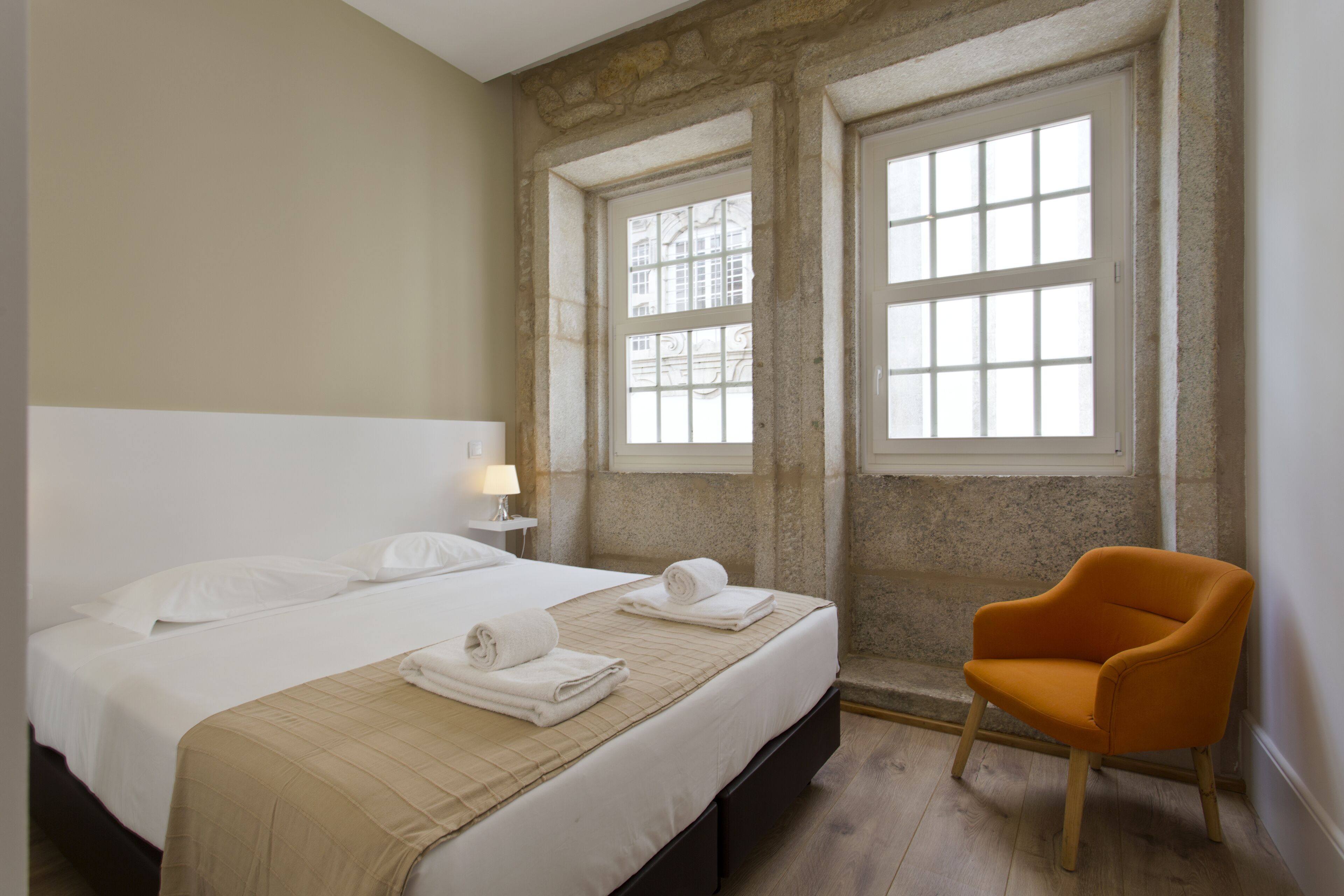 A hotel room with a bed, two windows, a chair, and a nightstand with a lamp.
