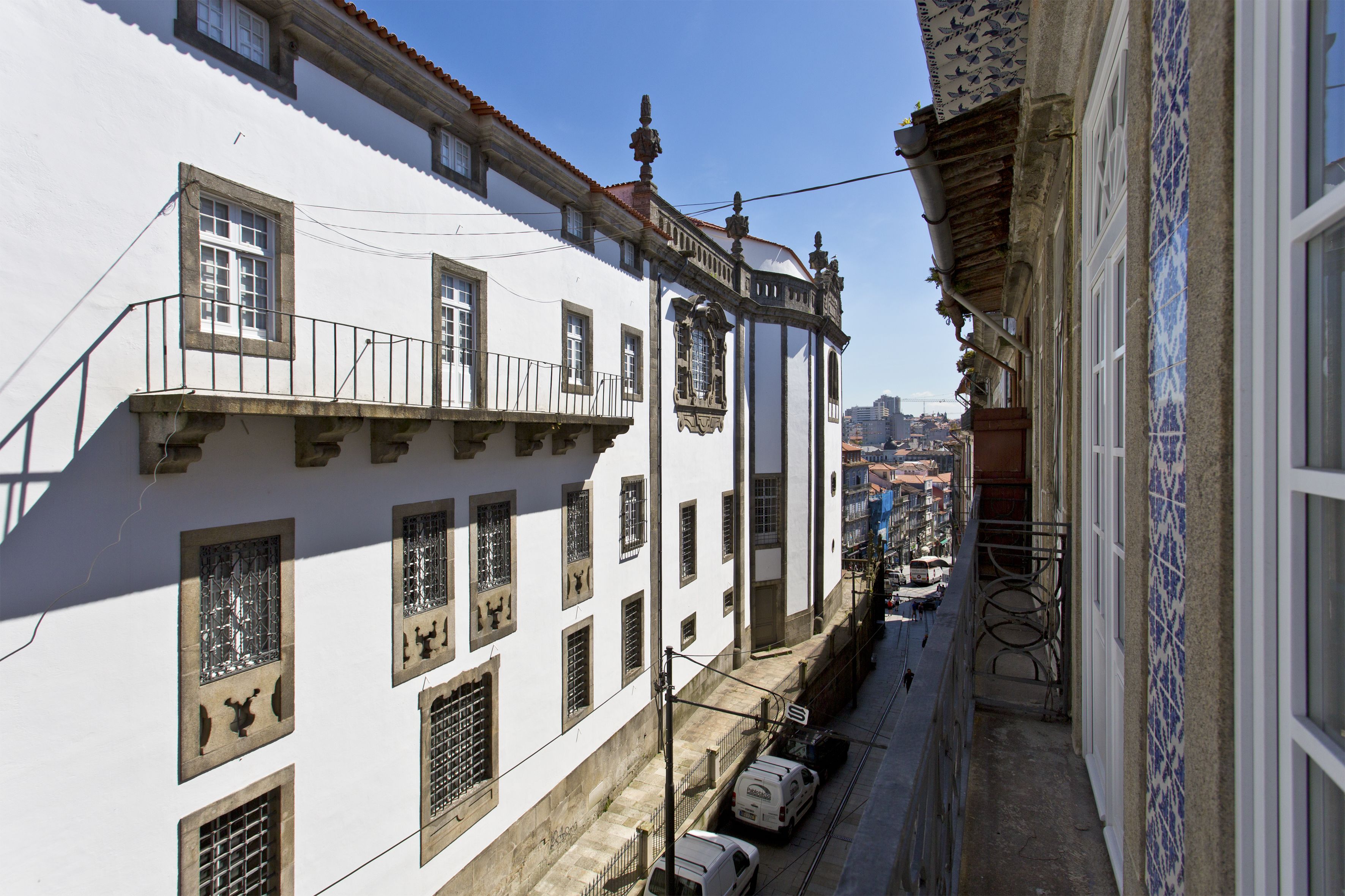A narrow street with white buildings, a tiled balcony, and a clear view of the city in the distance.