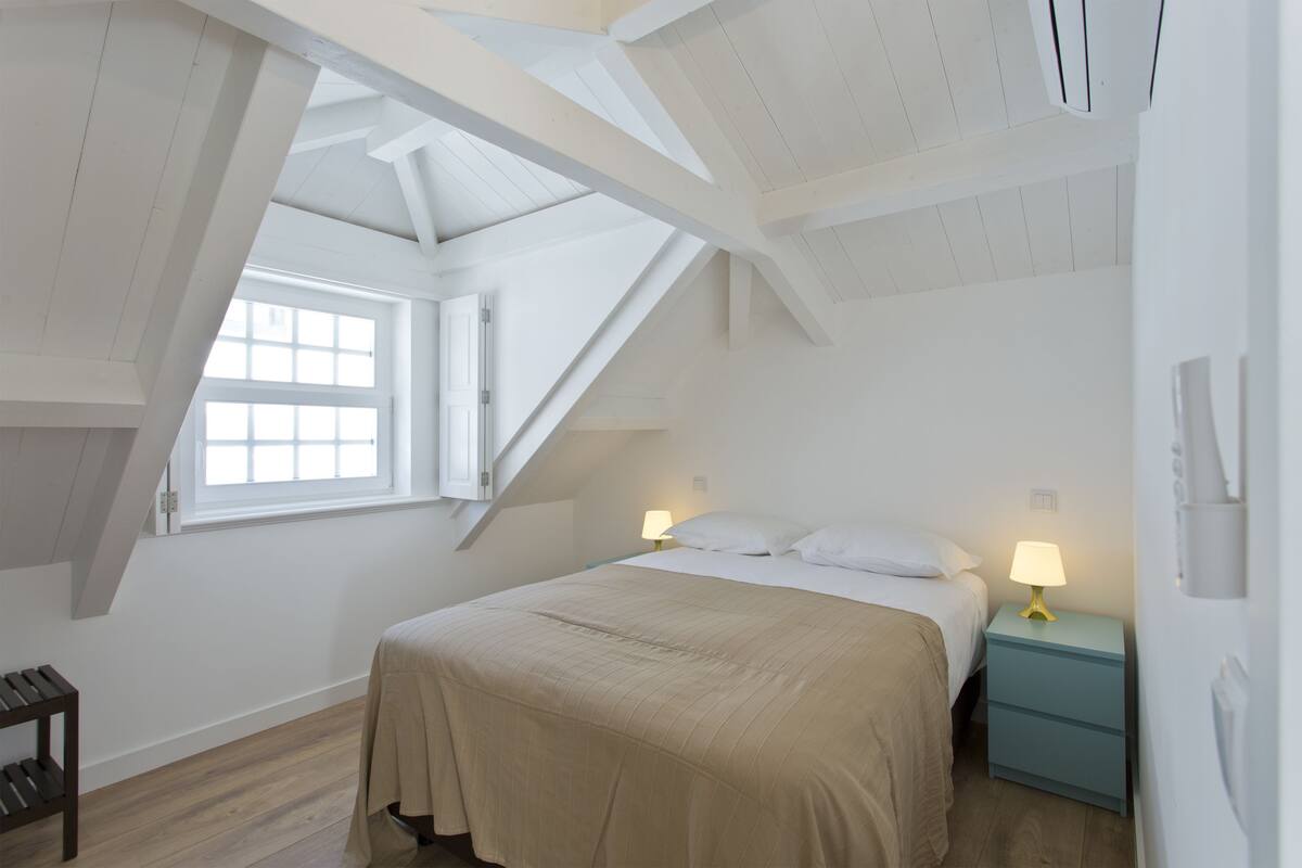 A bedroom with a sloped ceiling, a bed with white bedding, a nightstand with a lamp, and a window providing natural light.