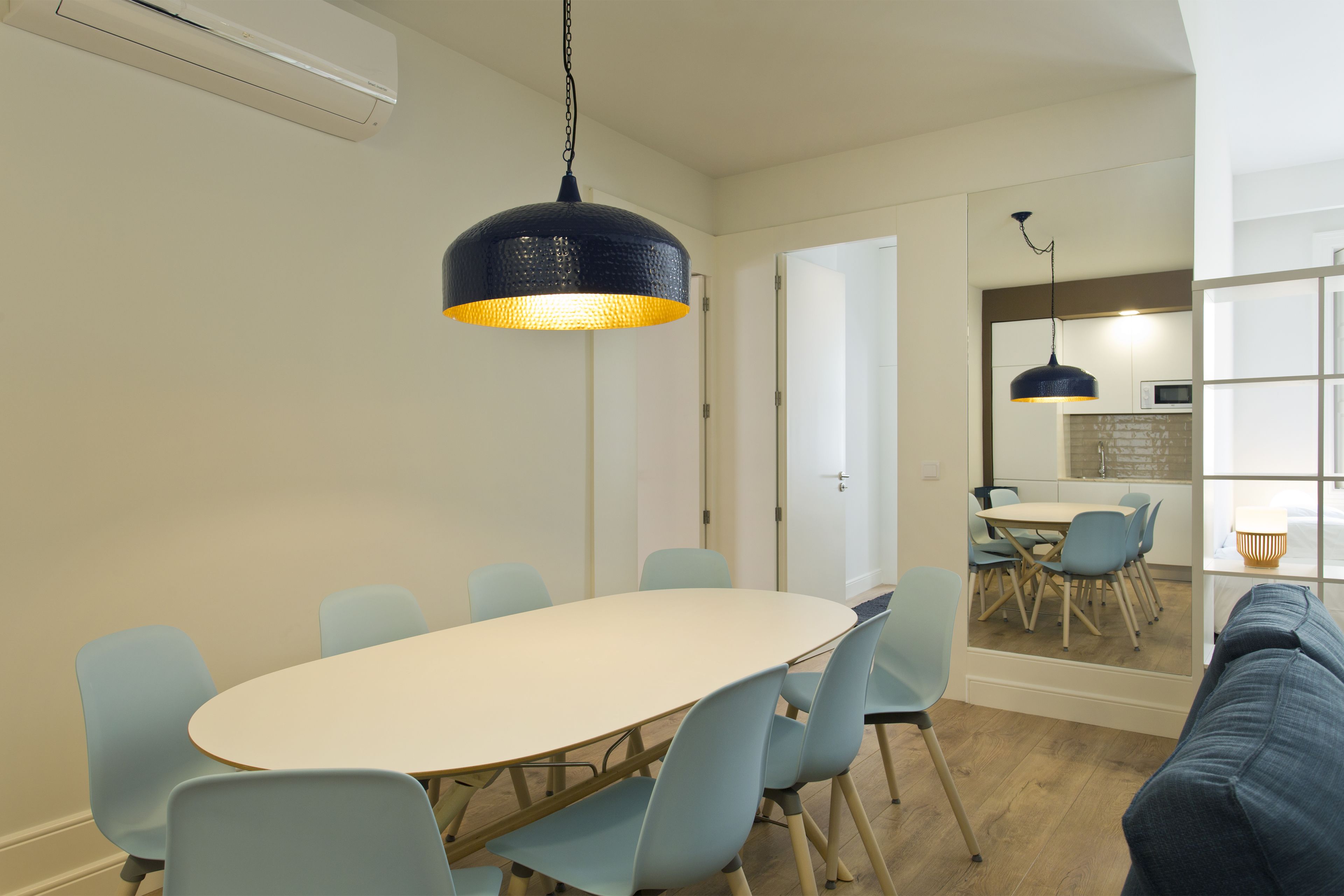 Un coin repas avec une table ronde et des chaises bleu clair, un luminaire suspendu bleu foncé, et une cuisine visible à travers une porte ouverte.