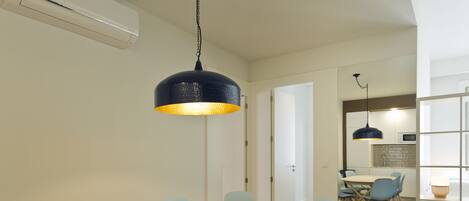 Un coin repas avec une table ronde et des chaises bleu clair, un luminaire suspendu bleu foncé, et une cuisine visible à travers une porte ouverte.