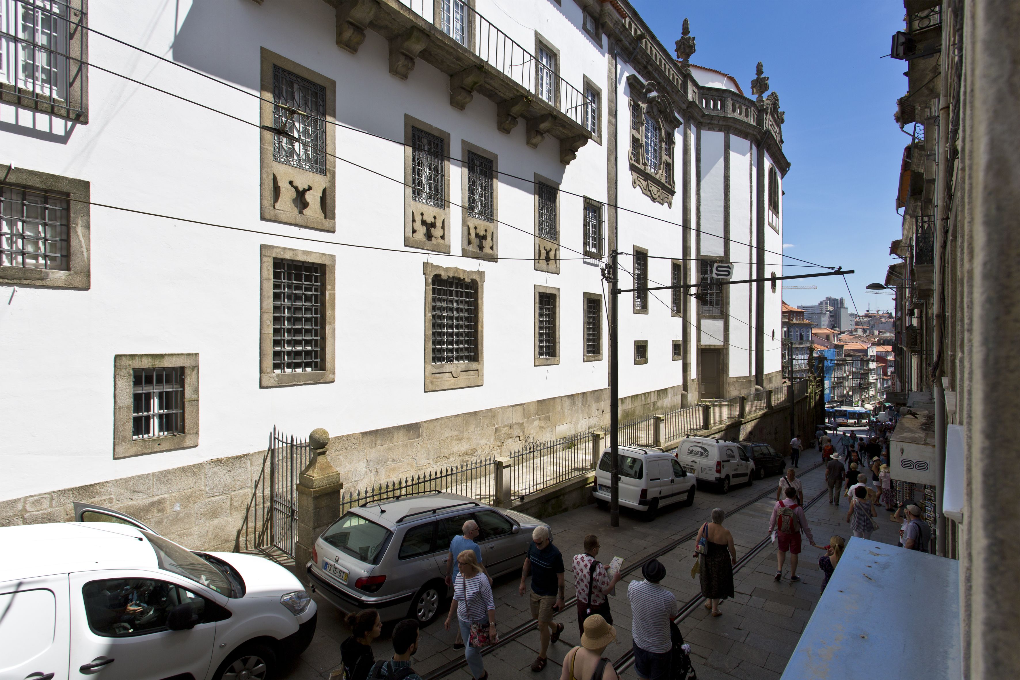 A narrow street with white buildings, a white van parked on the side, and pedestrians walking on the sidewalk.