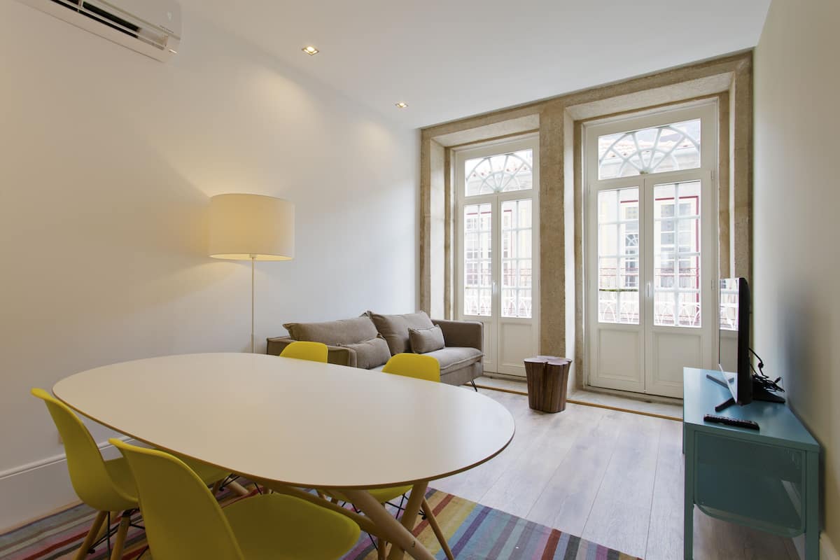 A room with a round table, yellow chairs, a grey sofa, a lamp, and a TV on a stand.