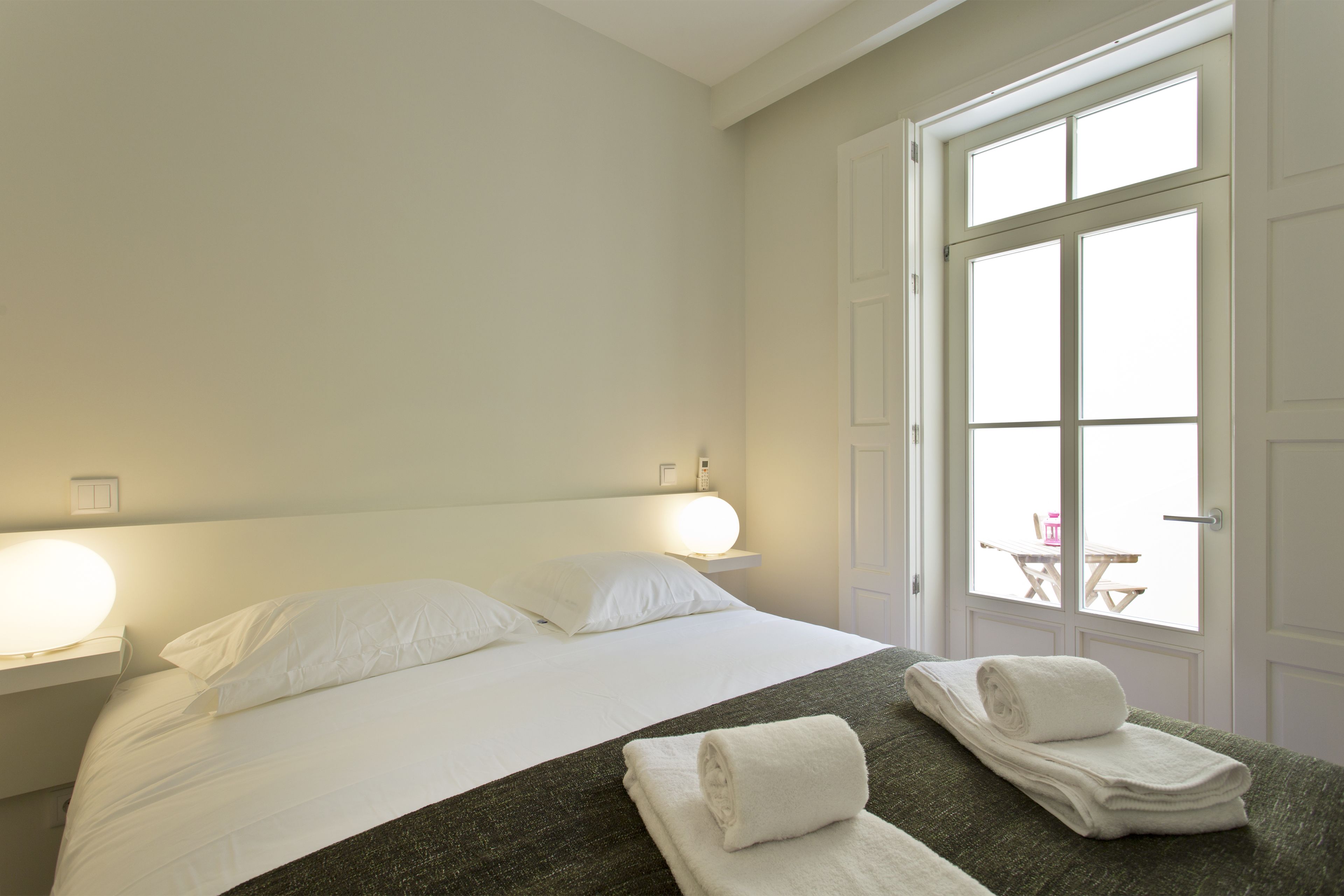 A hotel room with a bed, two bedside lamps, a window with a view, and a table with chairs outside.