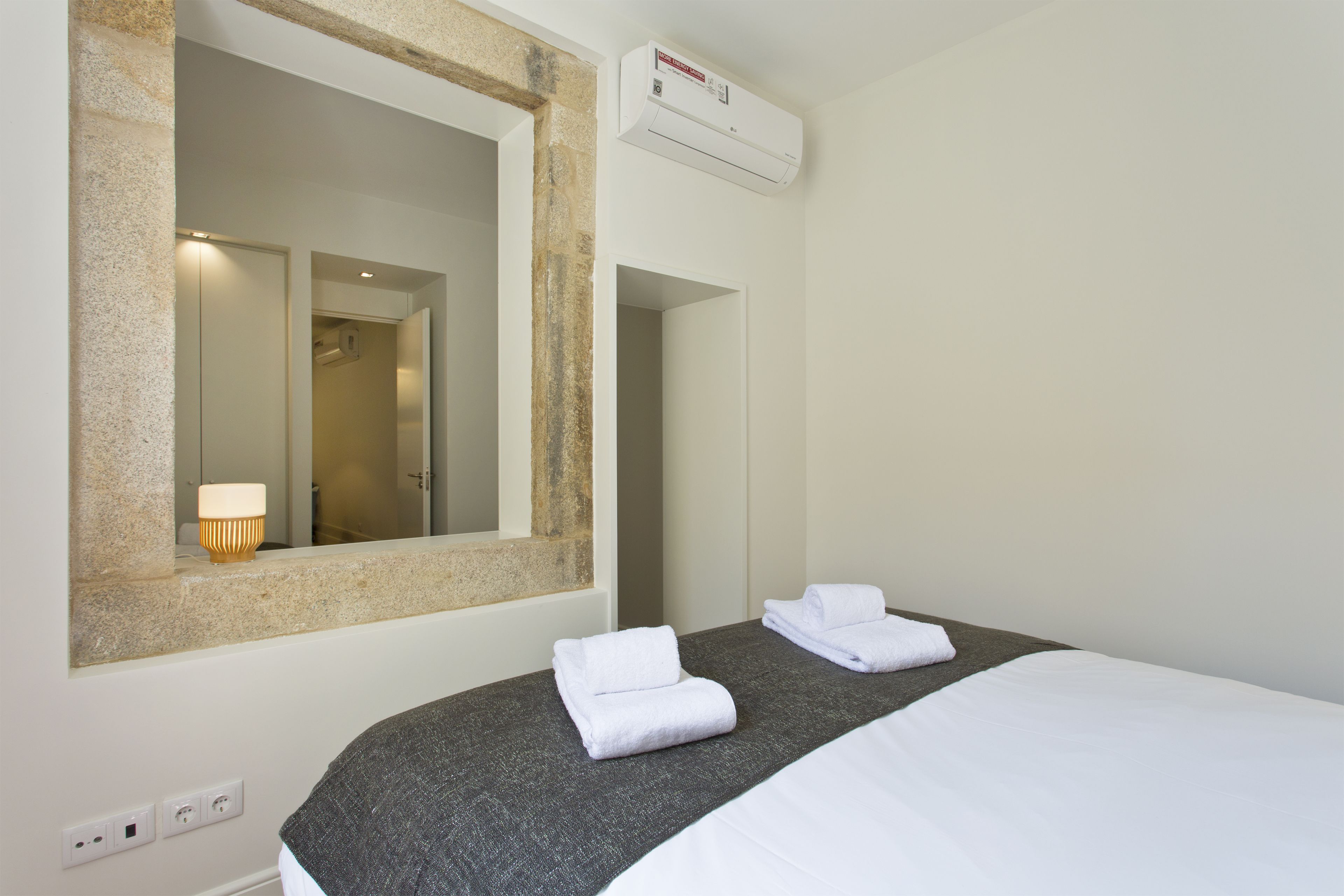 Une chambre d’hôtel avec un grand miroir, deux serviettes soigneusement pliées, un lit avec une tête de lit grise et un appareil de climatisation placé au-dessus du miroir.