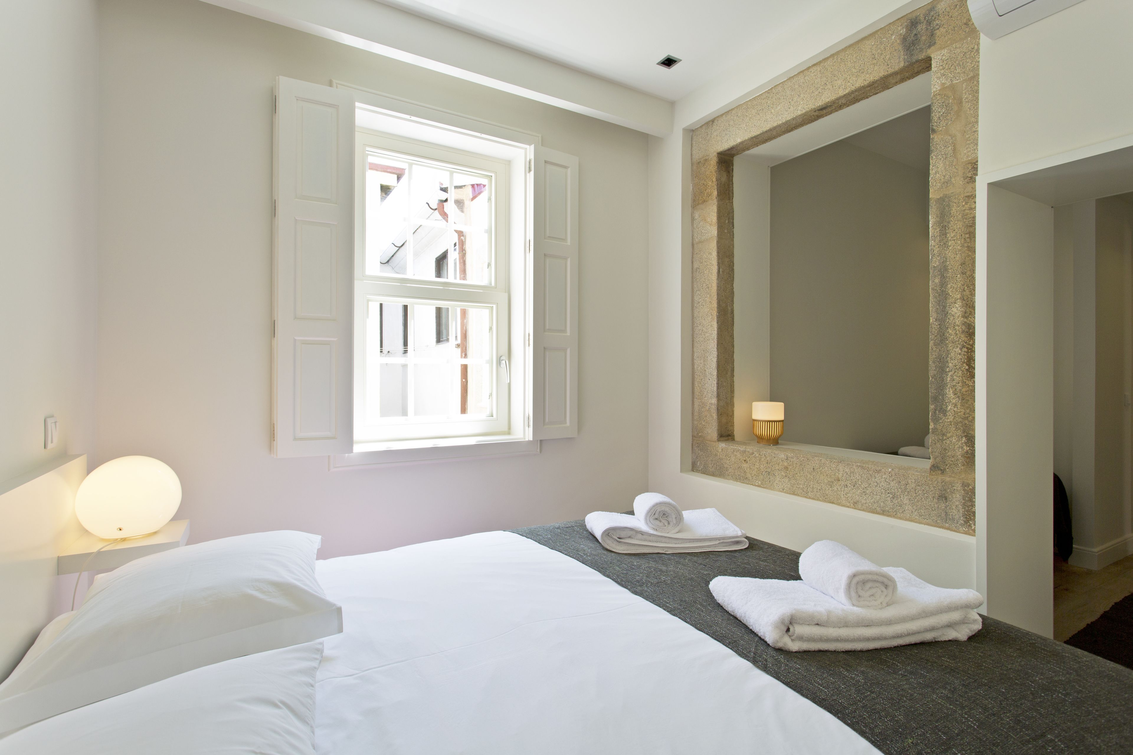 A hotel room with a bed, a window, a mirror, and a bedside table with a lamp.