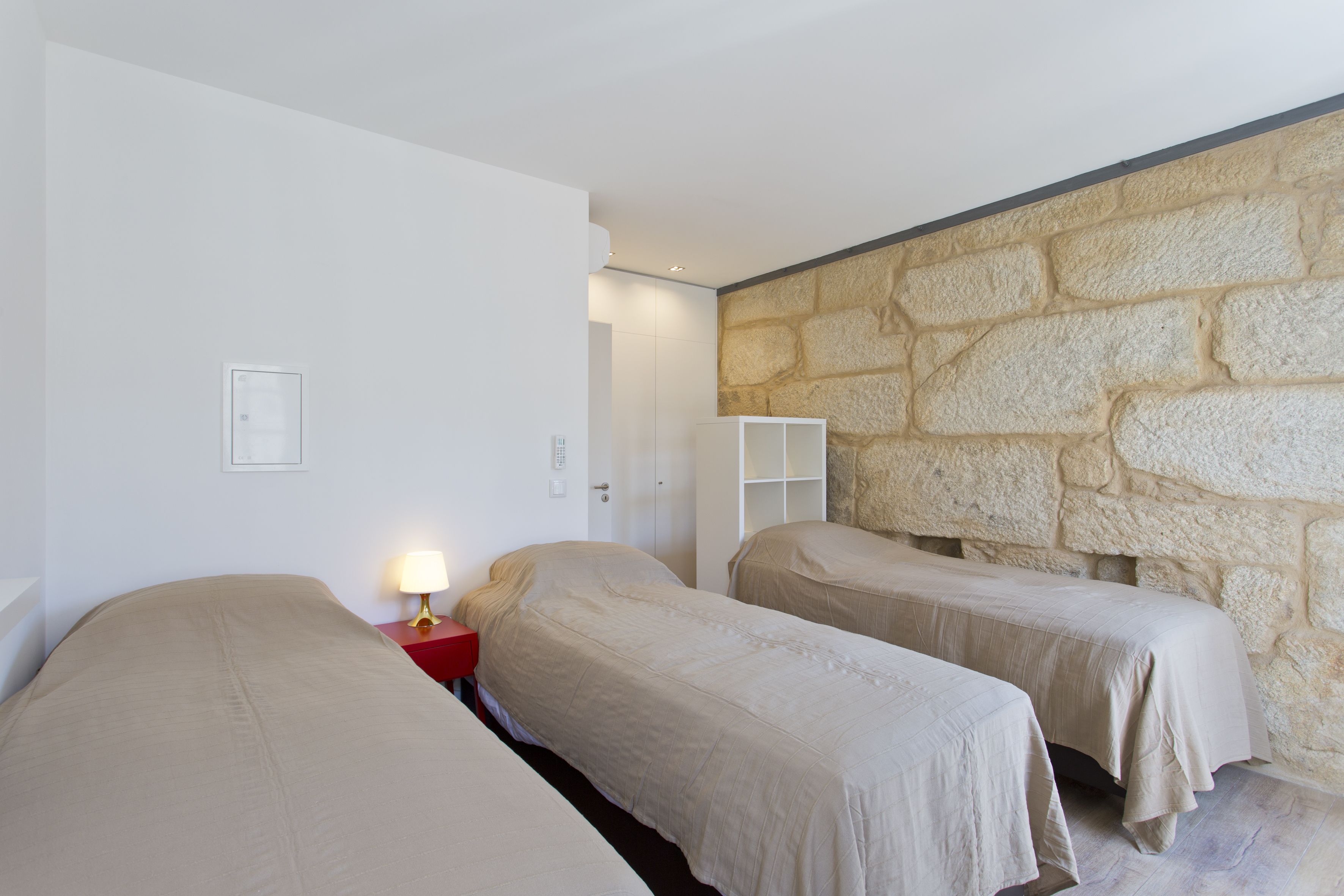 A bedroom with two beds, a red nightstand with a lamp, a white wardrobe, and a stone wall.