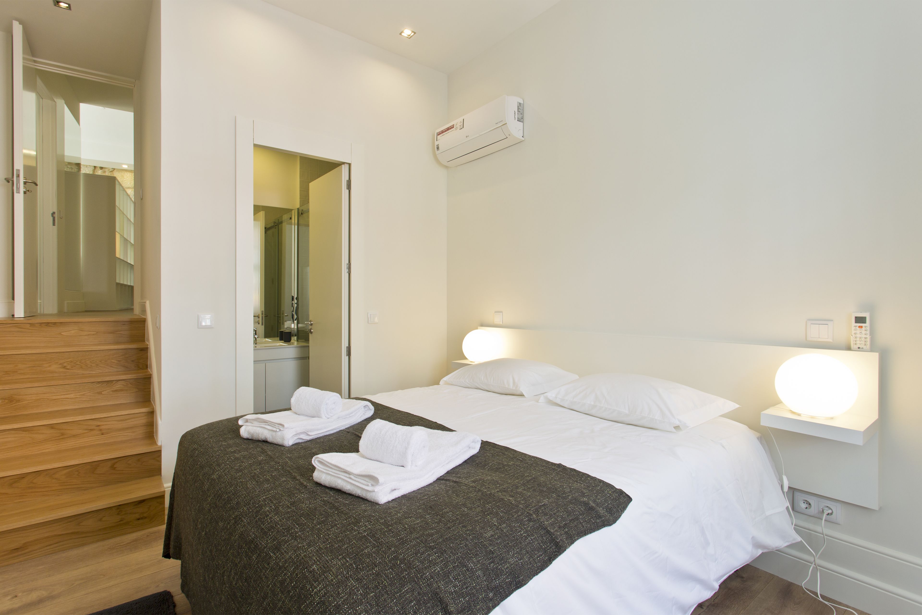Une chambre d’hôtel comprenant un lit, une table de chevet avec une lampe, un climatiseur fixé au mur et un escalier menant à une autre pièce.