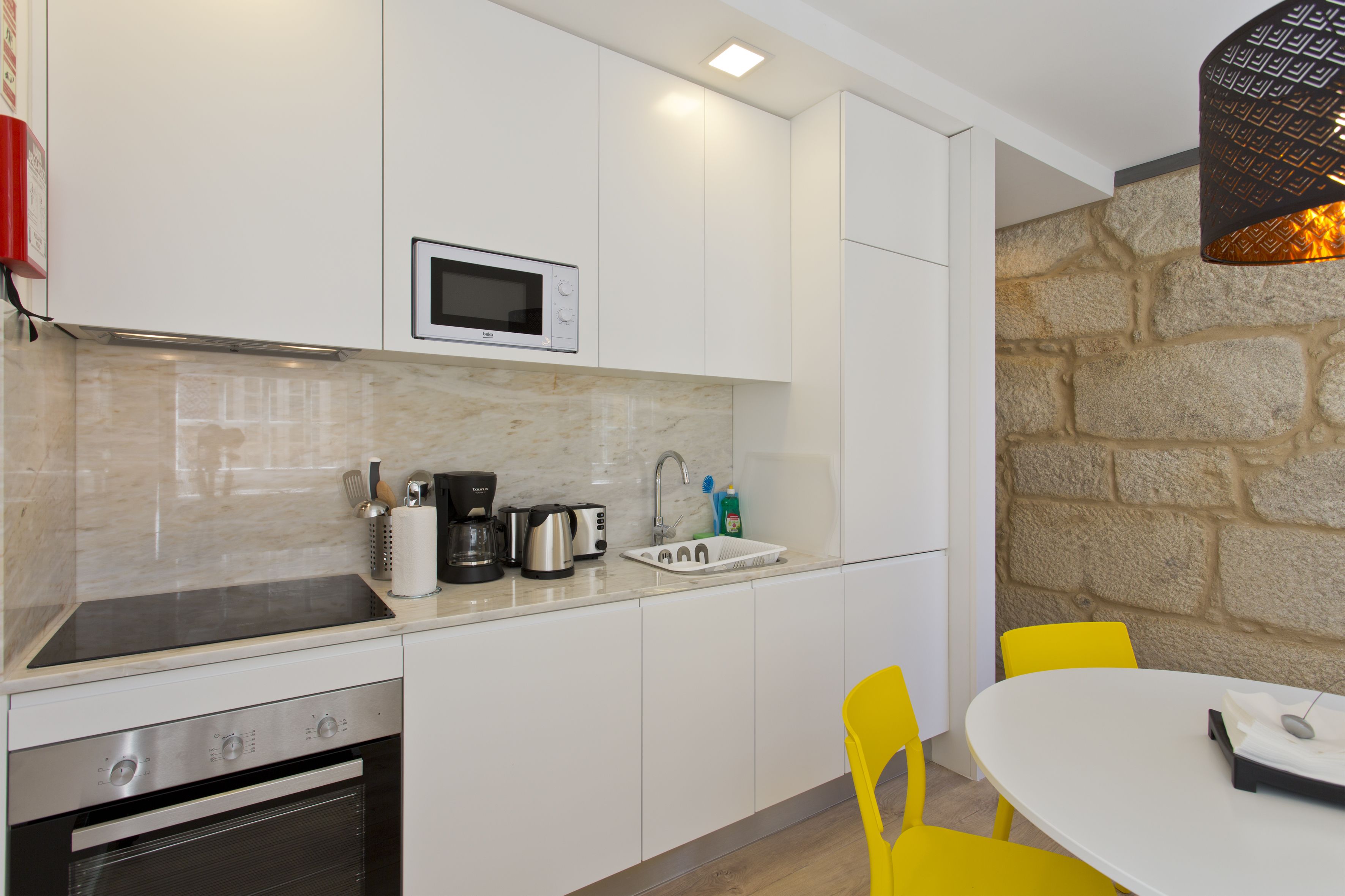 A compact kitchen with white cabinets, a microwave, a stove, and a sink. There is a stone wall and a table with chairs.