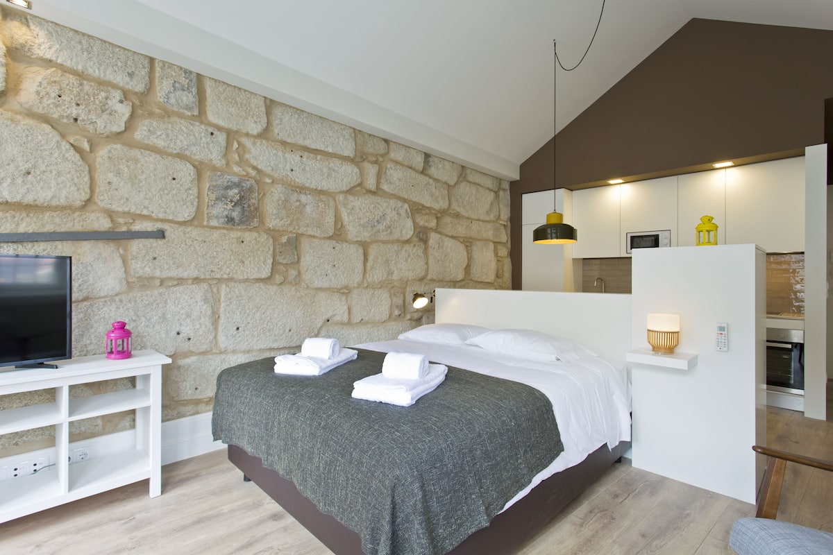 A modern bedroom with a stone wall, a bed with white linens, a TV, and a small table with a lamp.