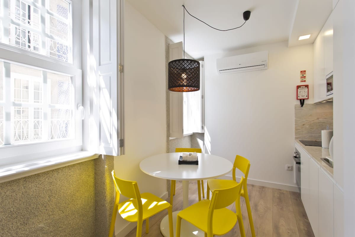 A dining area with a round table and four yellow chairs, a hanging pendant light, and a window with patterned glass.