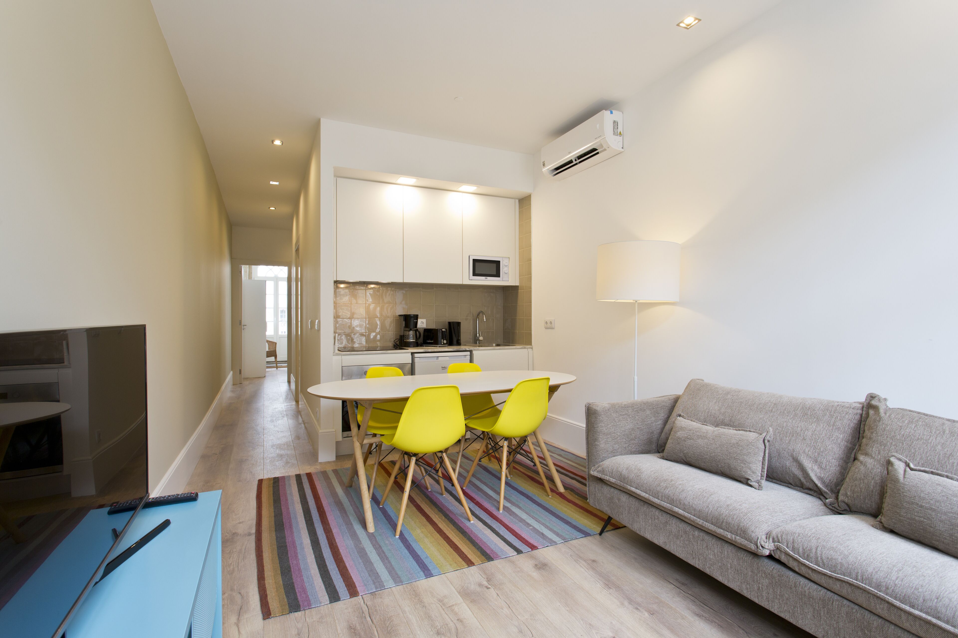 A modern living room with a grey sofa, a striped rug, and a dining area with yellow chairs.