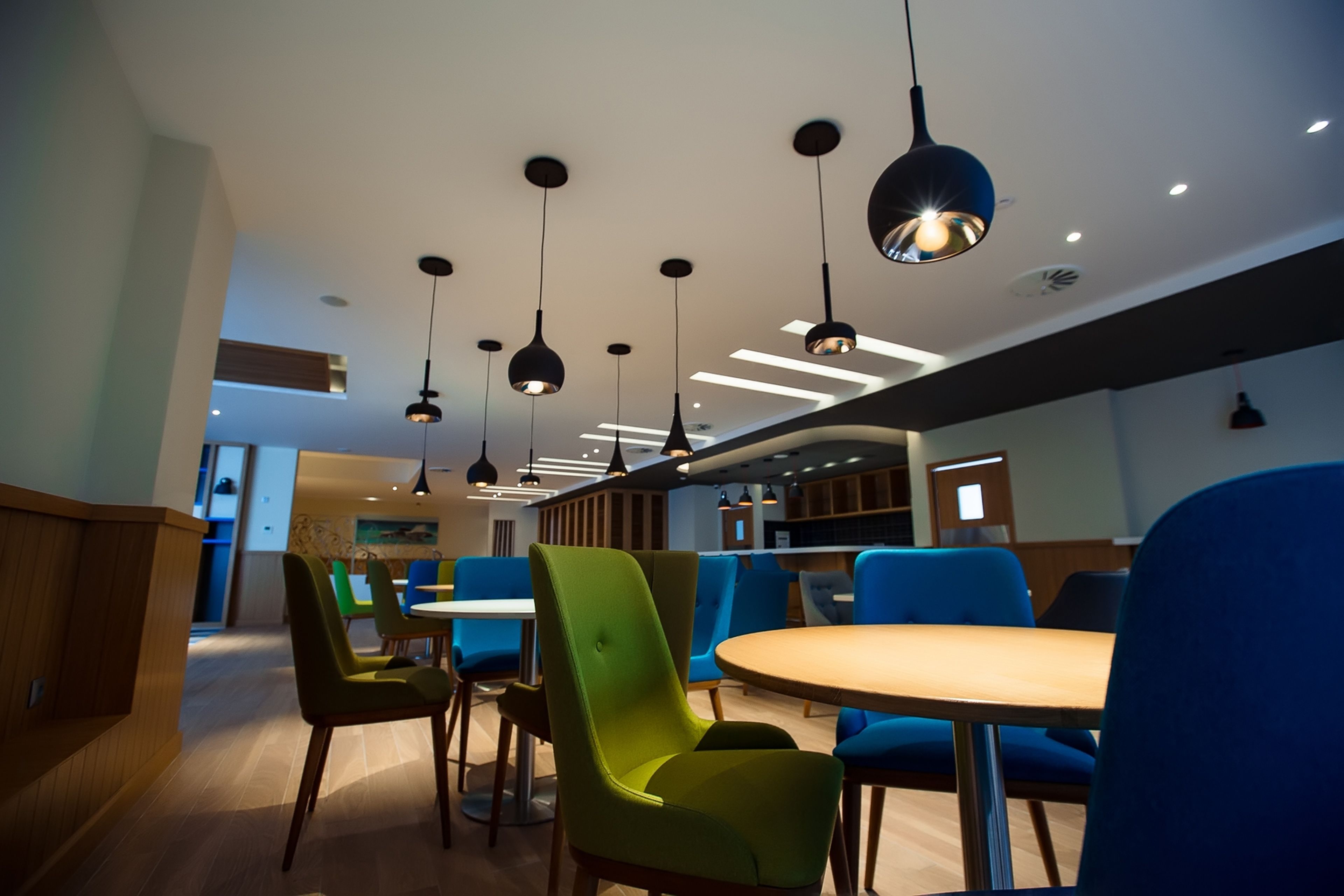 Un comedor moderno con sillas verdes y azules, mesas de madera e iluminación colgante.