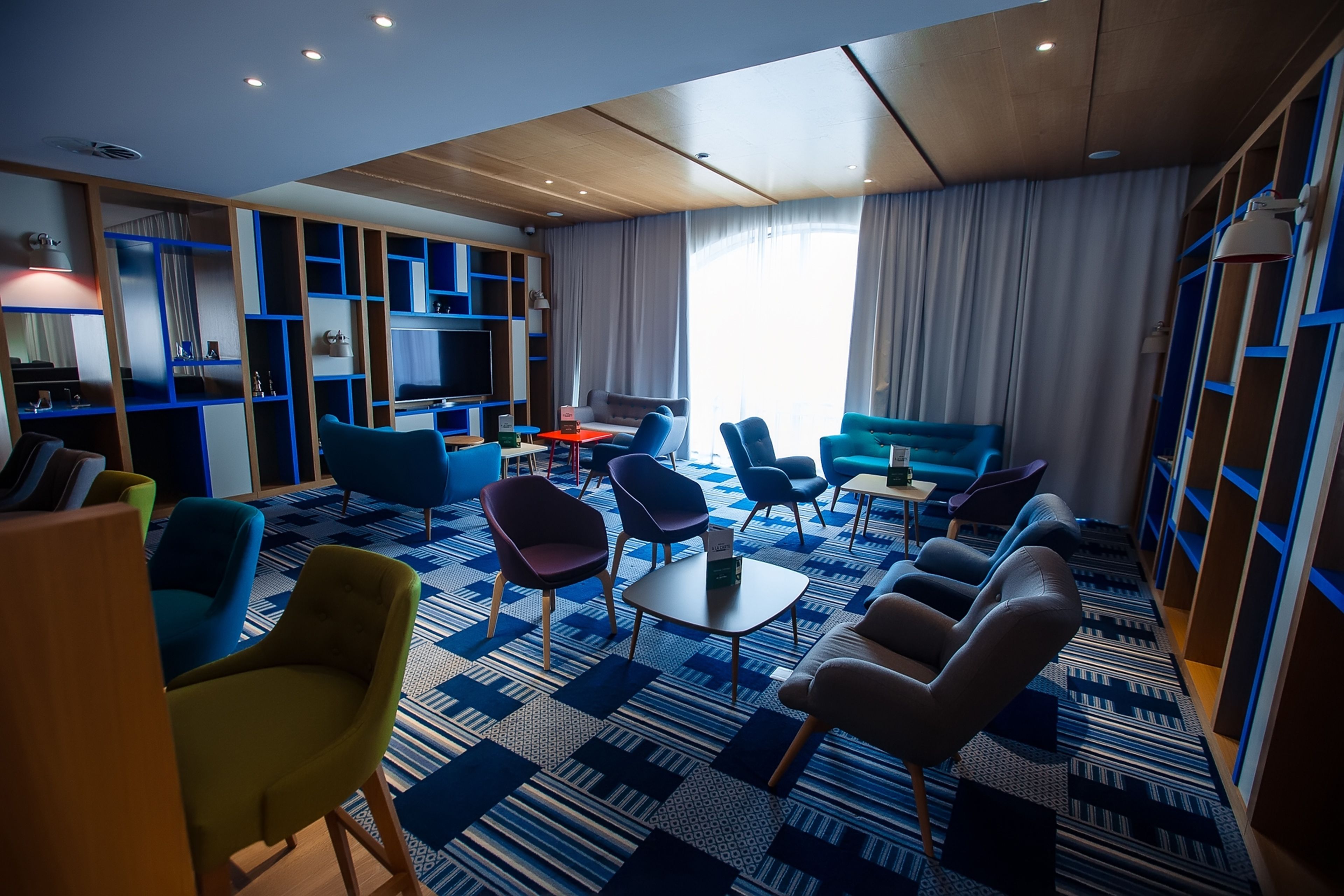 Un moderno vestíbulo de hotel con alfombra estampada, varias áreas de estar con sofás y sillones, un televisor y estanterías empotradas.