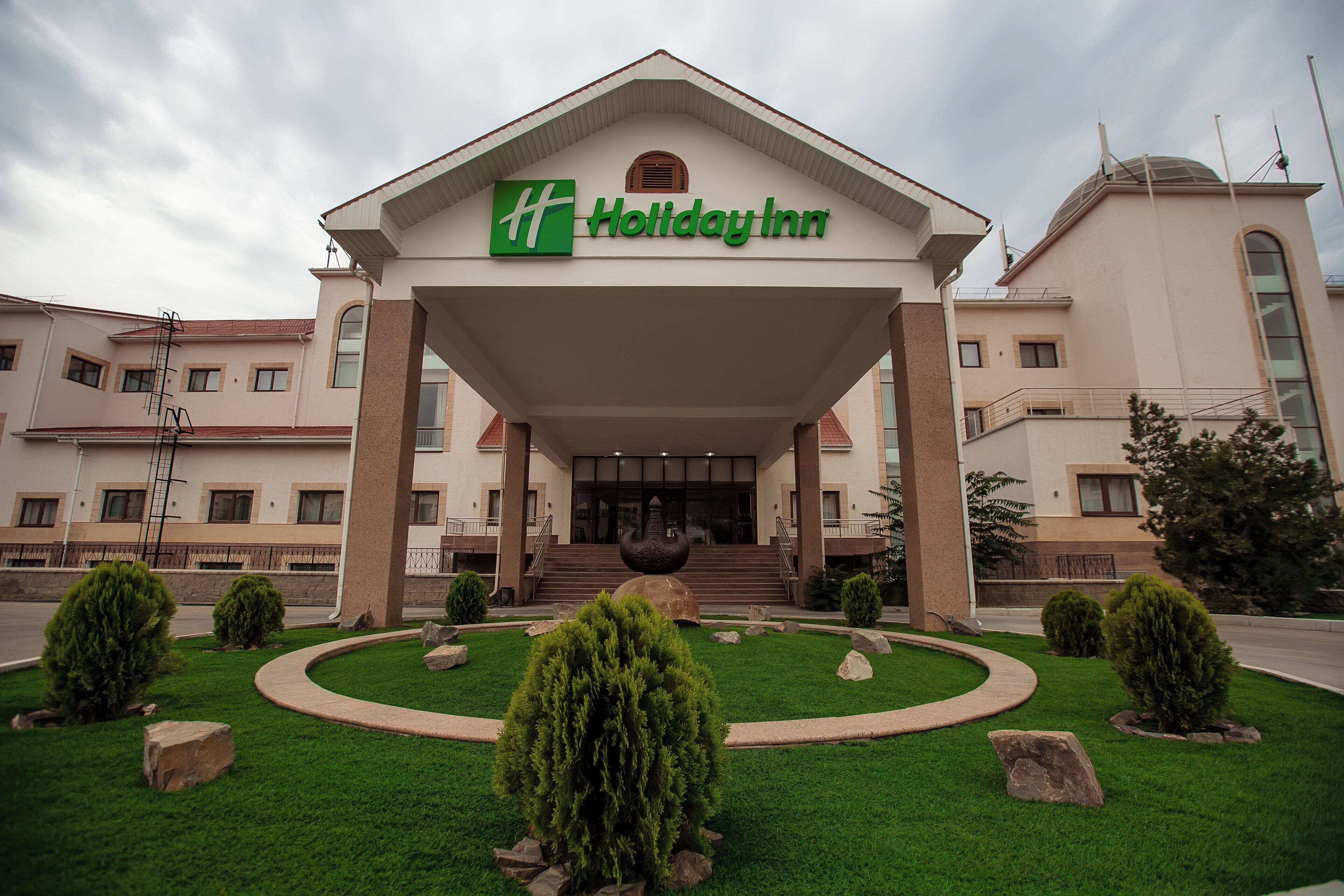 Entrada de un hotel Holiday Inn con un paseo techado, escalones que conducen a la entrada y un jardín ajardinado con arbustos y rocas.