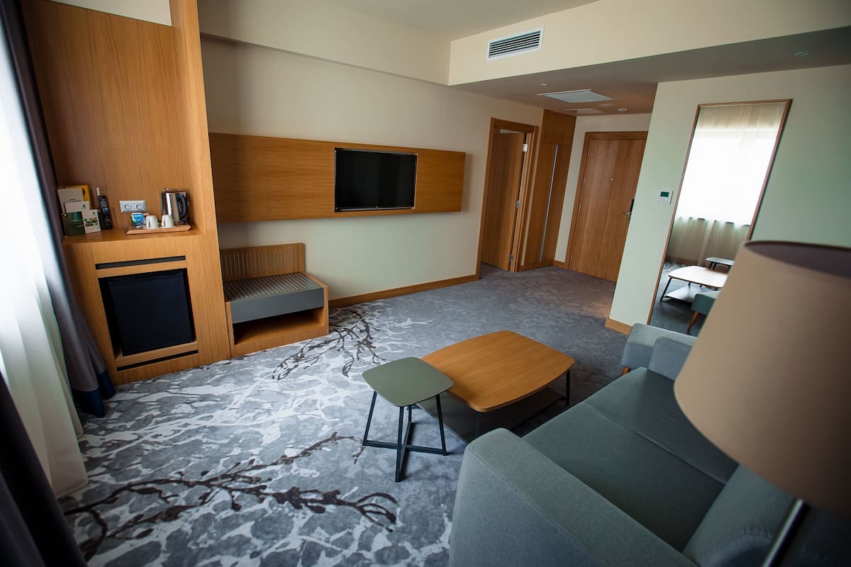 A modern hotel room with a sofa, a small table, a TV mounted on the wall, and a seating area with a bench.
