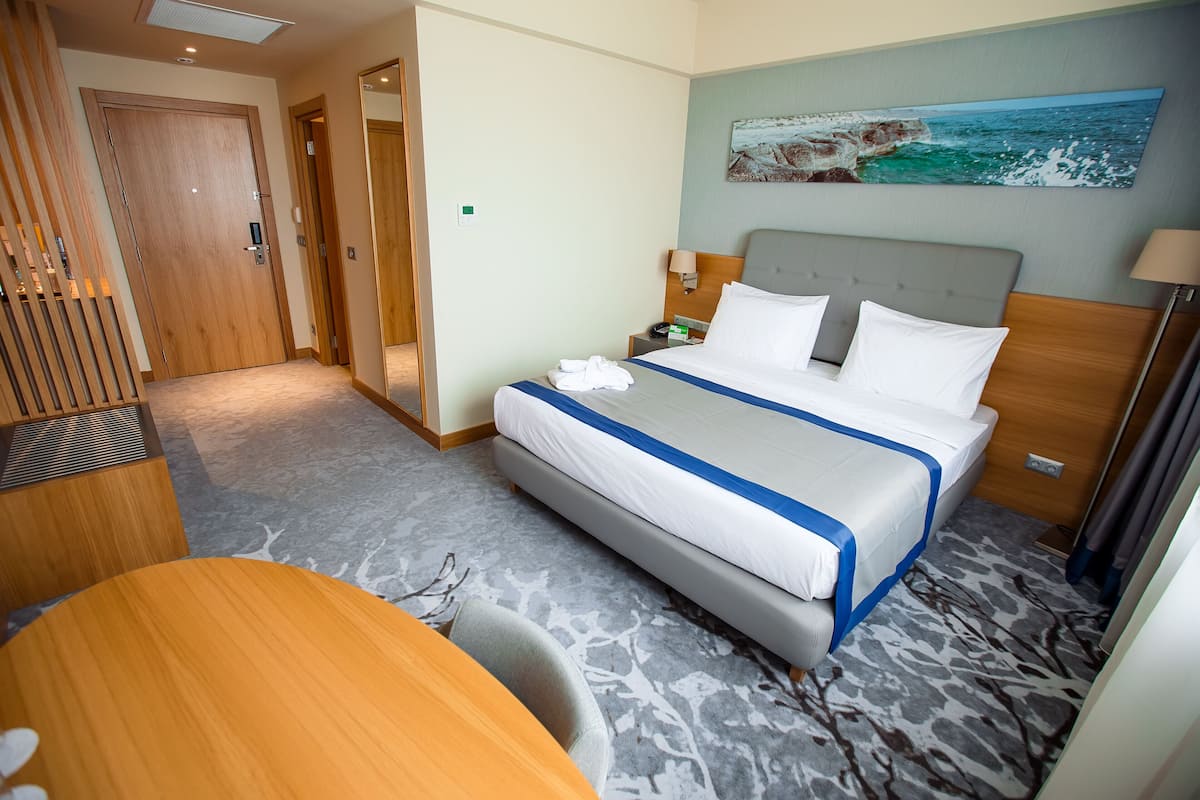 A hotel room with a bed, a desk, and a chair. There is a painting on the wall and a door in the background.