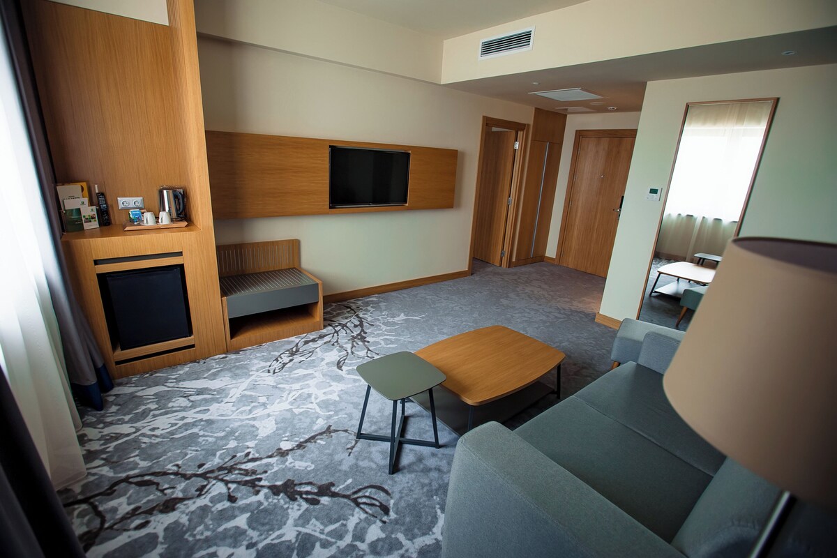 Une chambre d’hôtel moderne équipée d’un canapé, d’une petite table, d’un téléviseur fixé au mur et d’un meuble en bois contenant un téléviseur à écran plat.