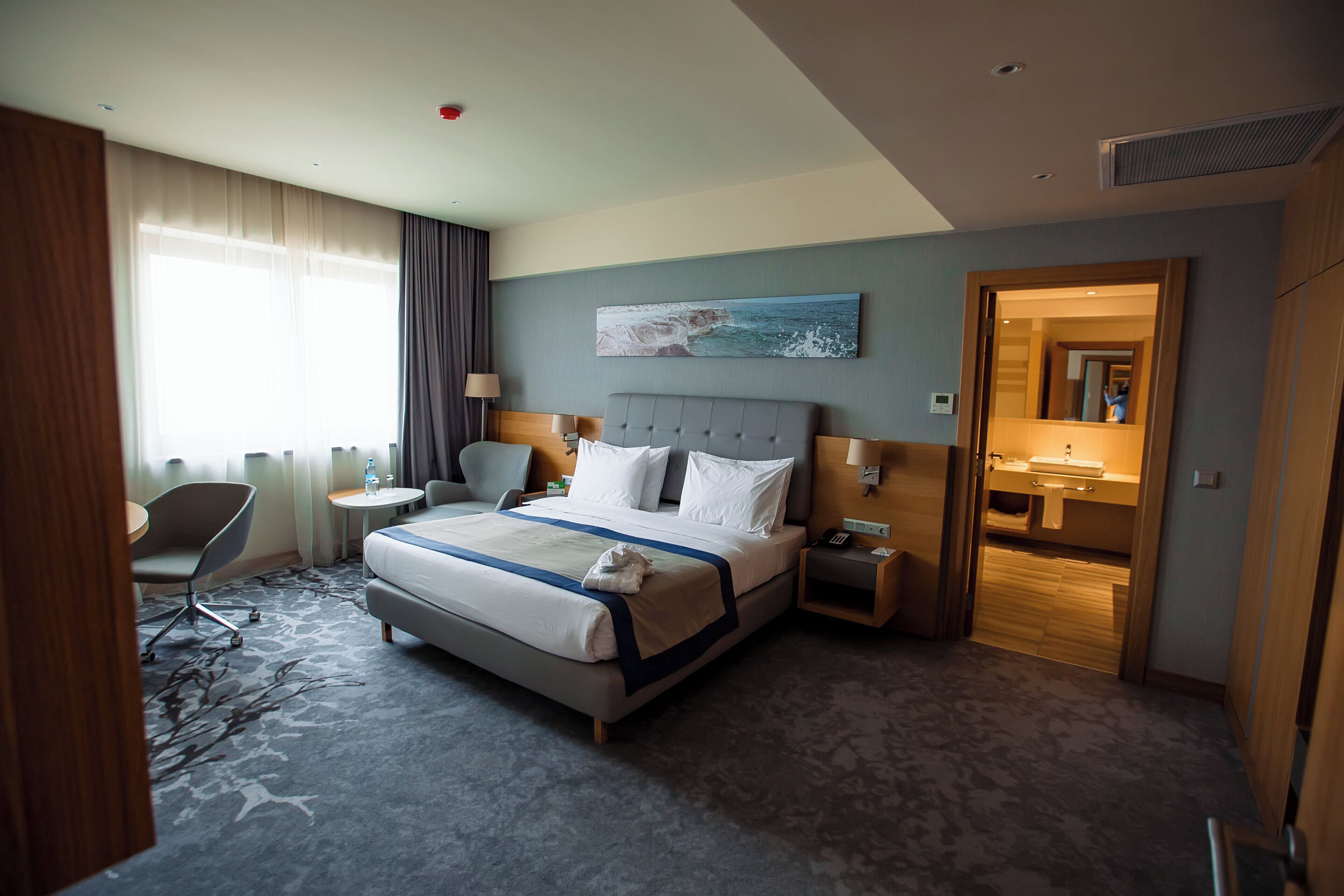 A hotel room with a large bed, a desk, a chair, a small table, a painting, and a bathroom visible through an open door.