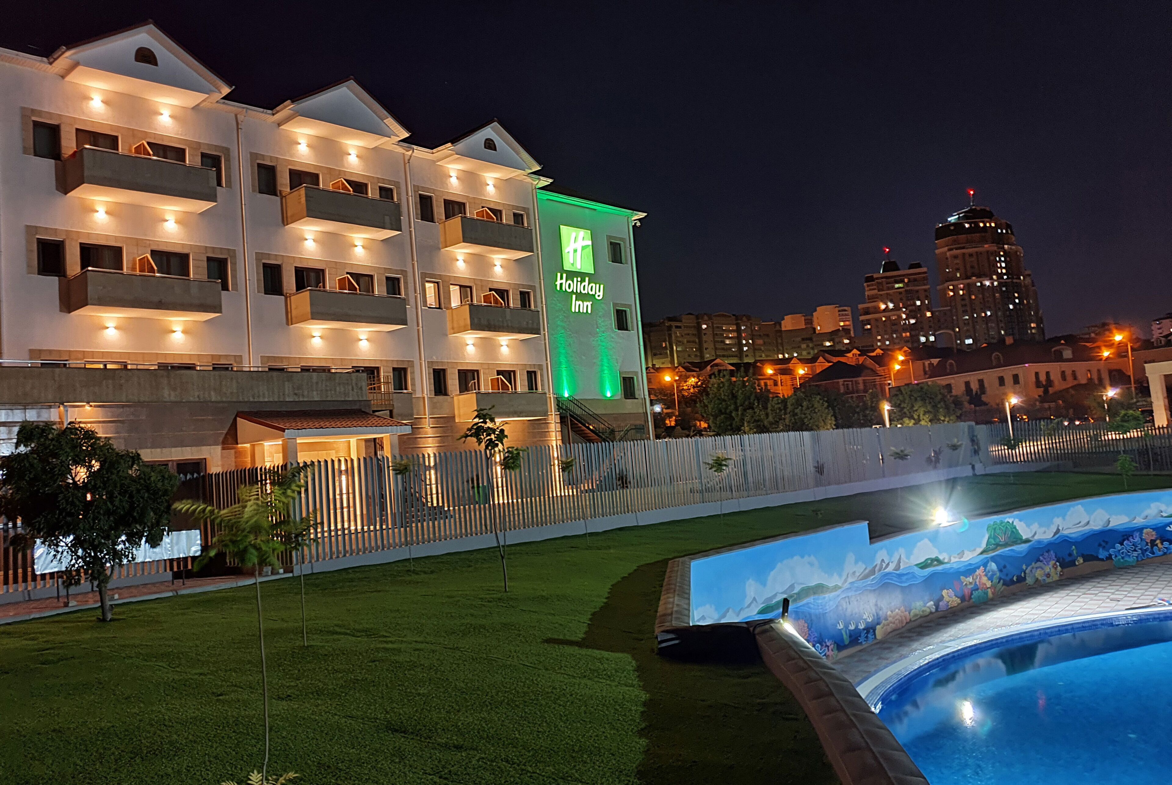 A hotel with multiple balconies, a swimming pool, and a city skyline in the background.
