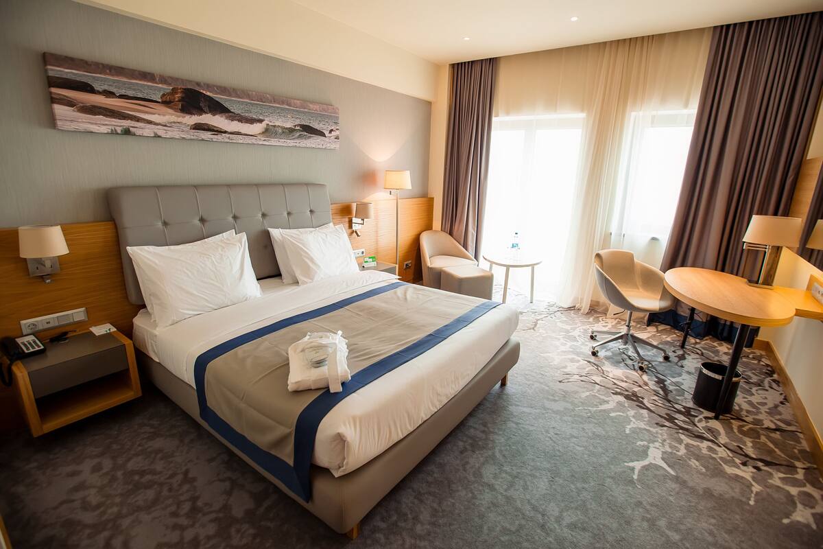A hotel room with a large bed, two bedside tables, a desk, a chair, and a view of the sea through the window.