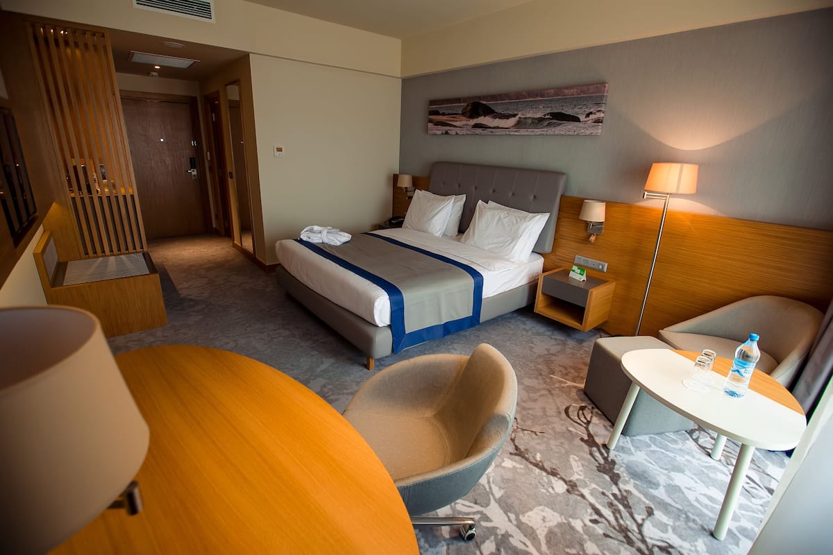 A hotel room with a bed, a desk, a chair, a small table, and a lamp.