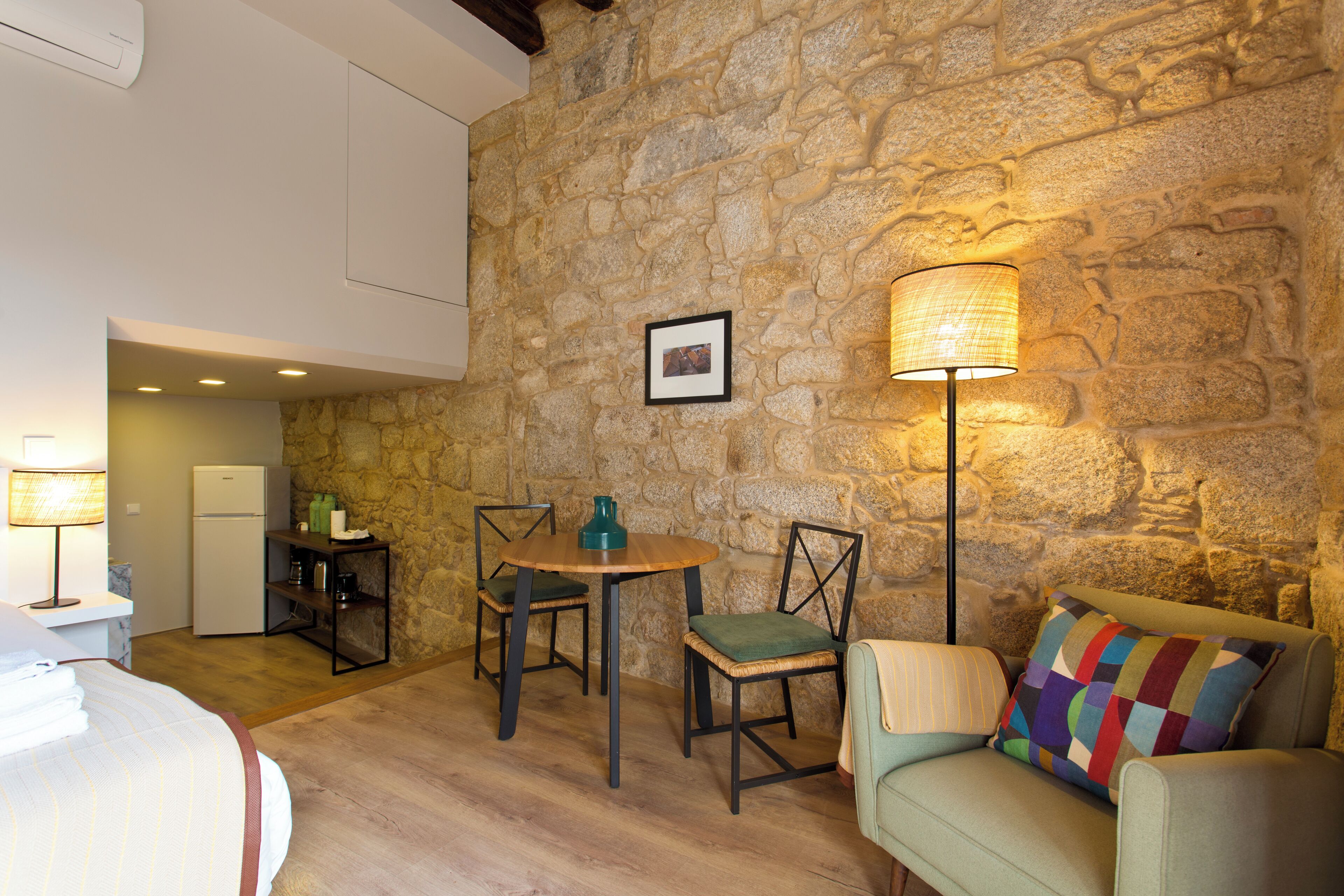 A cozy room with a stone wall, a wooden table with chairs, a sofa with a colorful pillow, and a lamp.