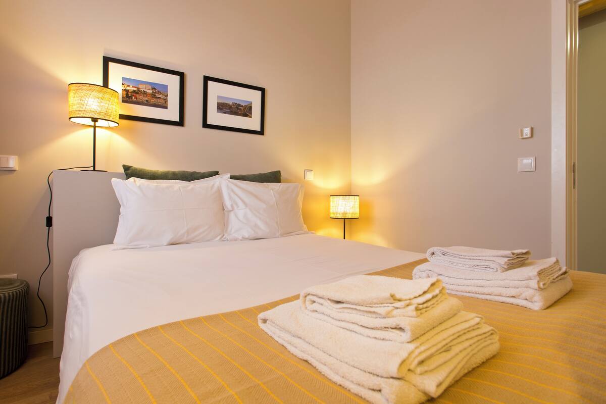 A hotel room with a bed, two wall-mounted lamps, a framed picture, and a stack of towels on the bed.