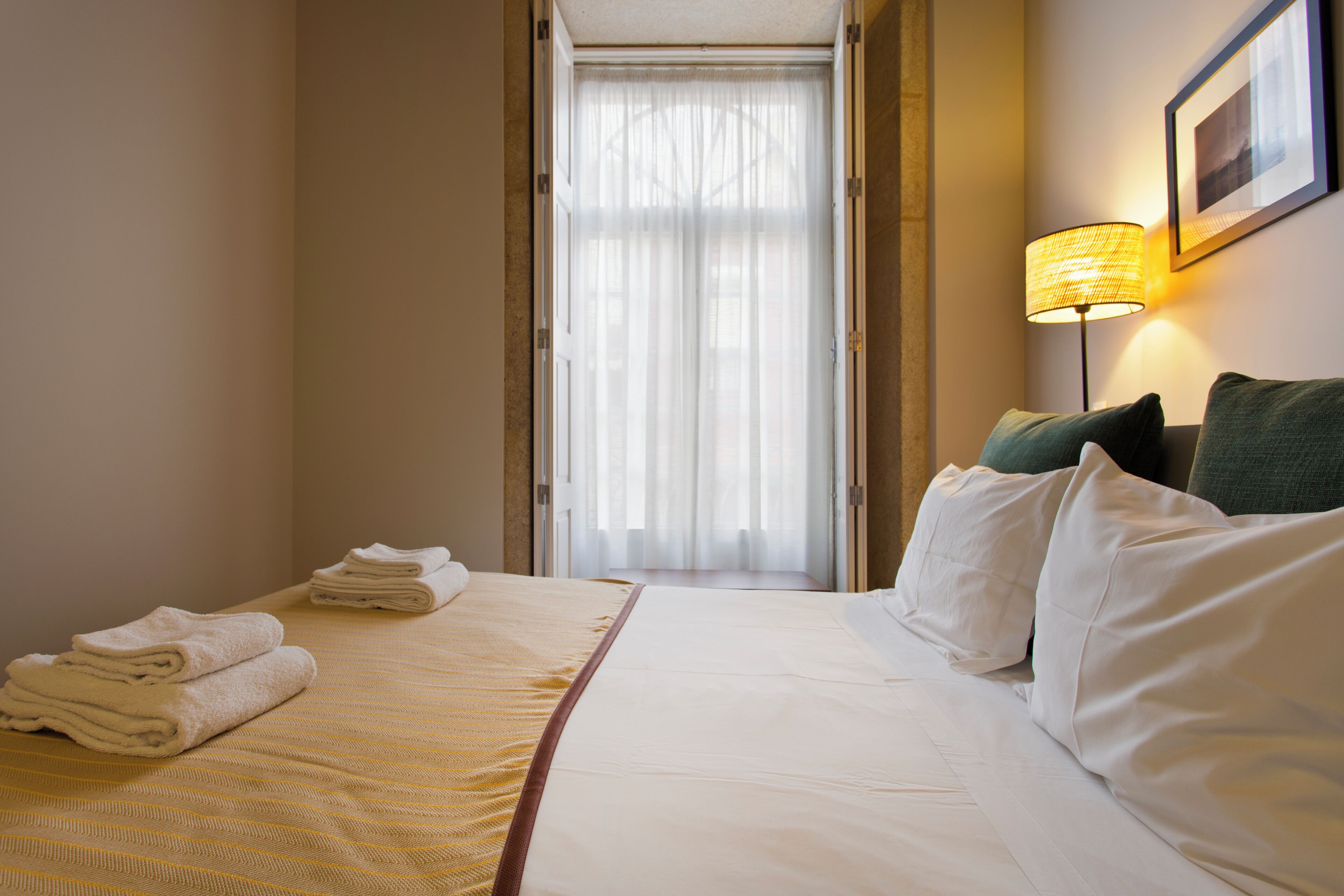 Una habitación de hotel con una cama bien hecha, una lámpara de pie y vistas a través de una ventana con cortinas de gasa.