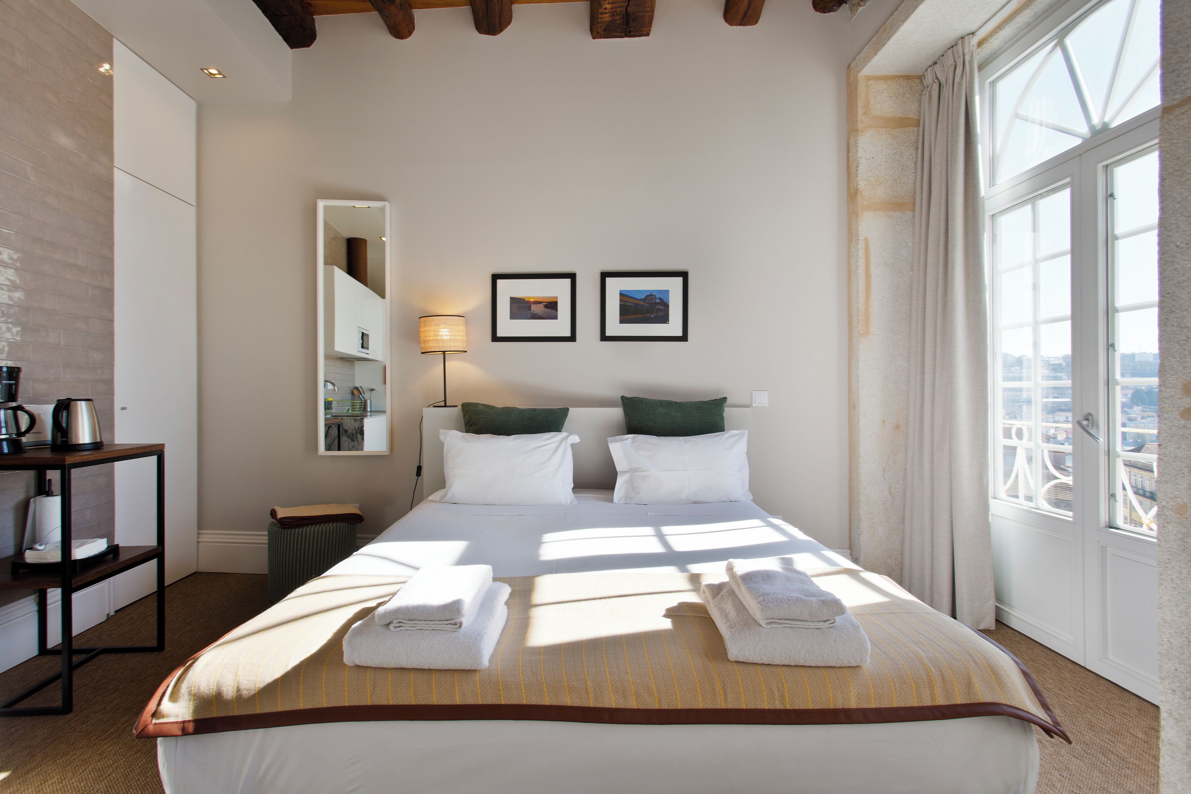 A hotel room with a large bed, two pillows, a bedside table with a lamp, and a view of the city through a large window.