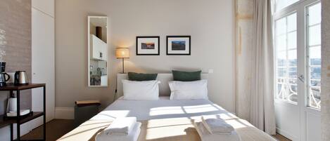 A hotel room with a large bed, two pillows, a bedside table with a lamp, and a view of the city through a large window.