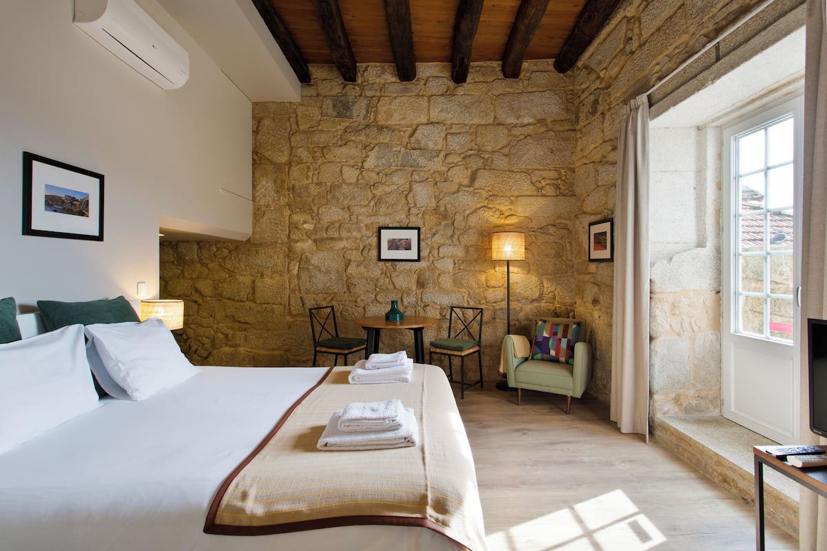 A hotel room with a stone wall, a bed with white linens, a small table with chairs, and a window with curtains.