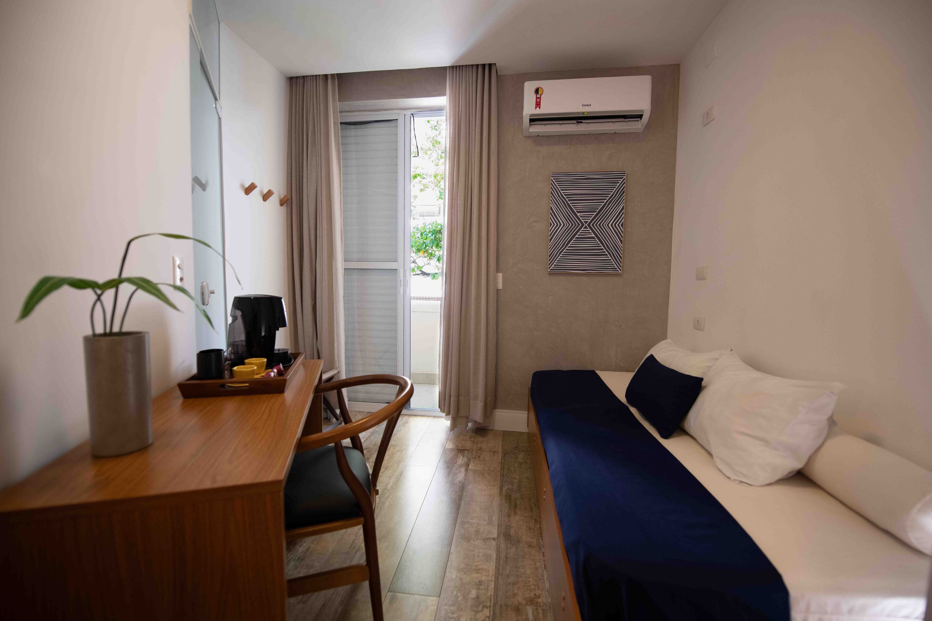 Una habitación compacta con una cama, un escritorio y una silla. Hay una planta en maceta sobre el escritorio y un equipo de aire acondicionado montado en la pared.