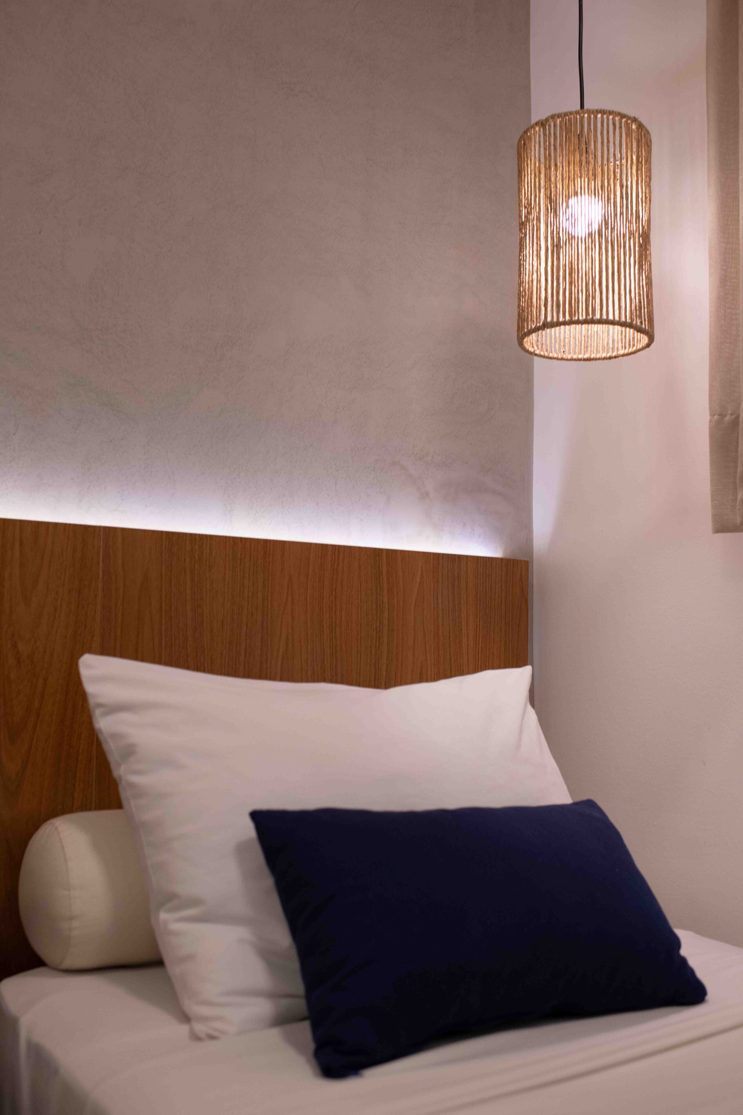 Un dormitorio con cabecera de madera, almohadas blancas y azul oscuro, y una lámpara colgante de fibras naturales.