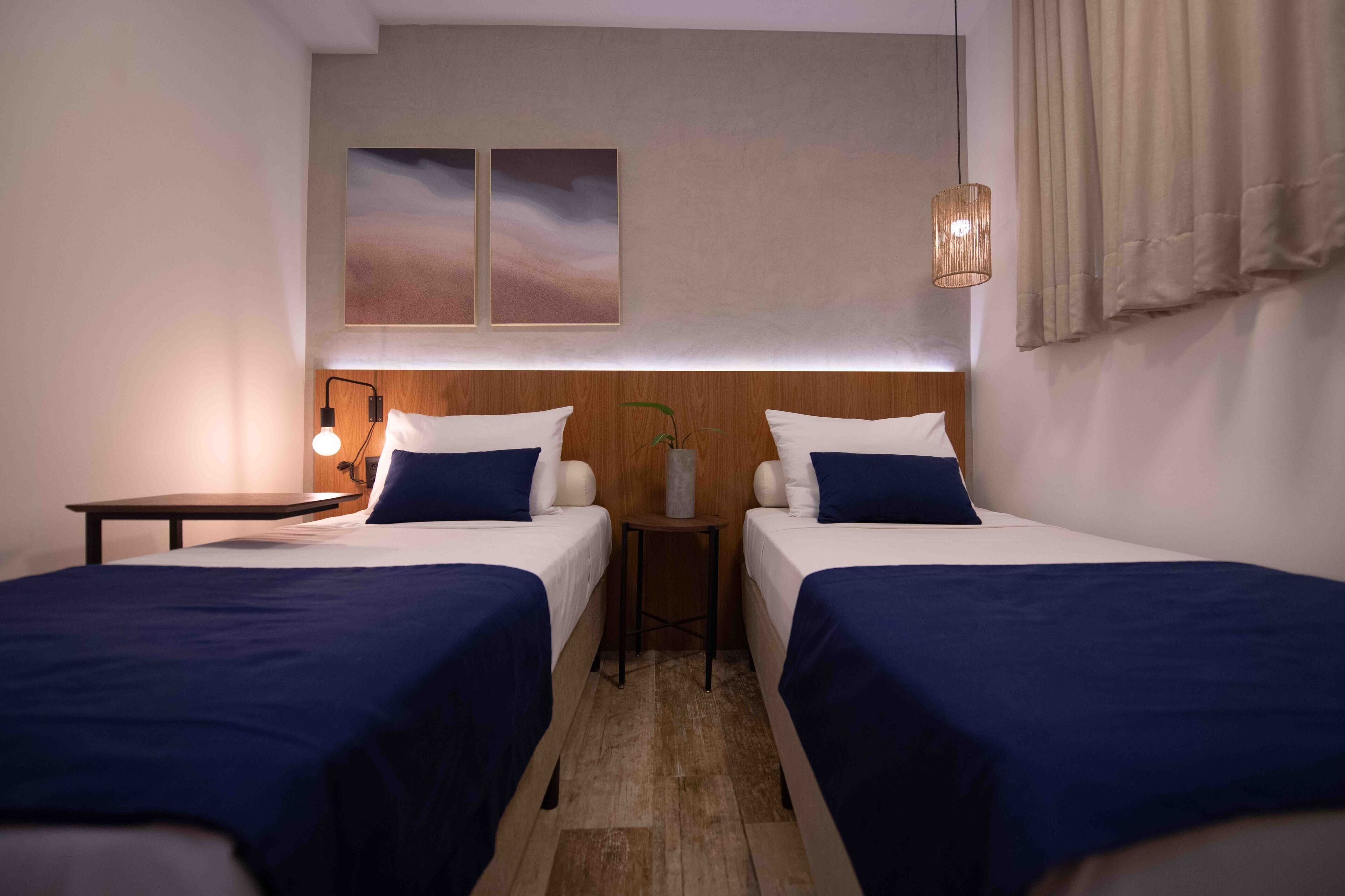 A hotel room with two beds, a small table, a lamp, and a wall-mounted artwork.