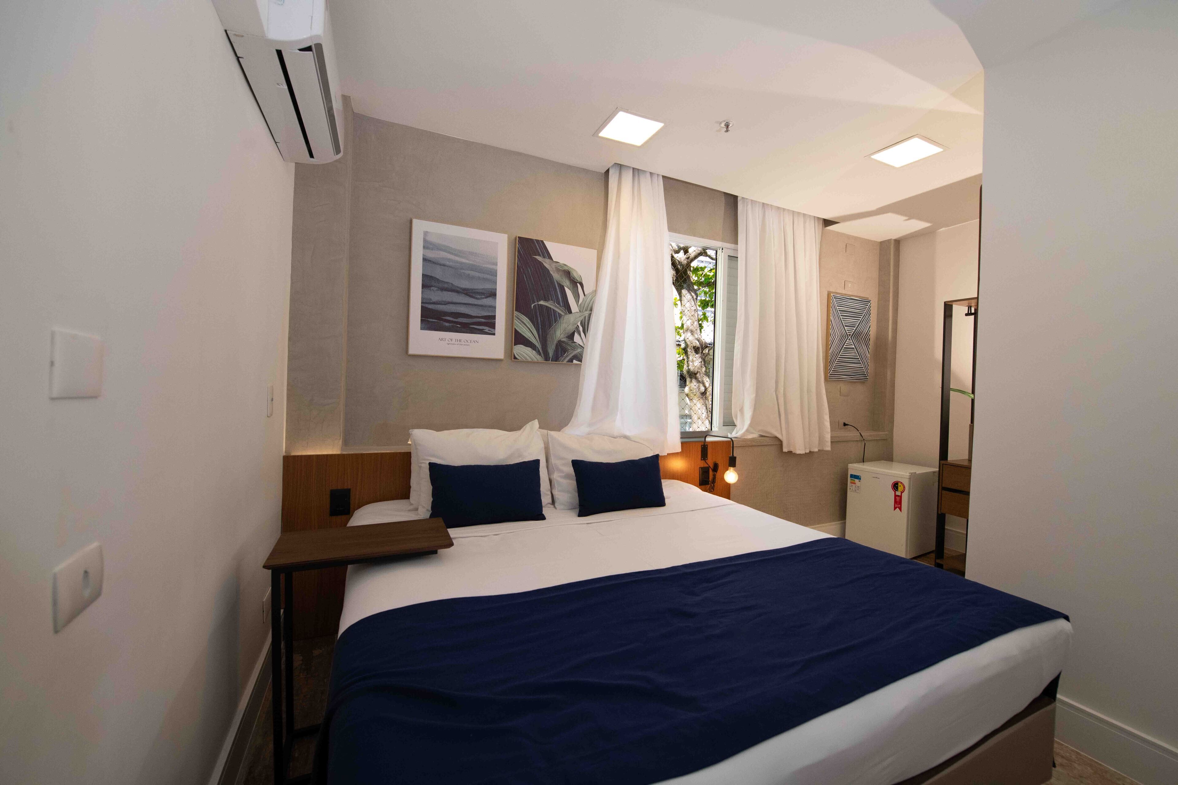 A hotel room with a bed, bedside table, air conditioning unit, and a small refrigerator.