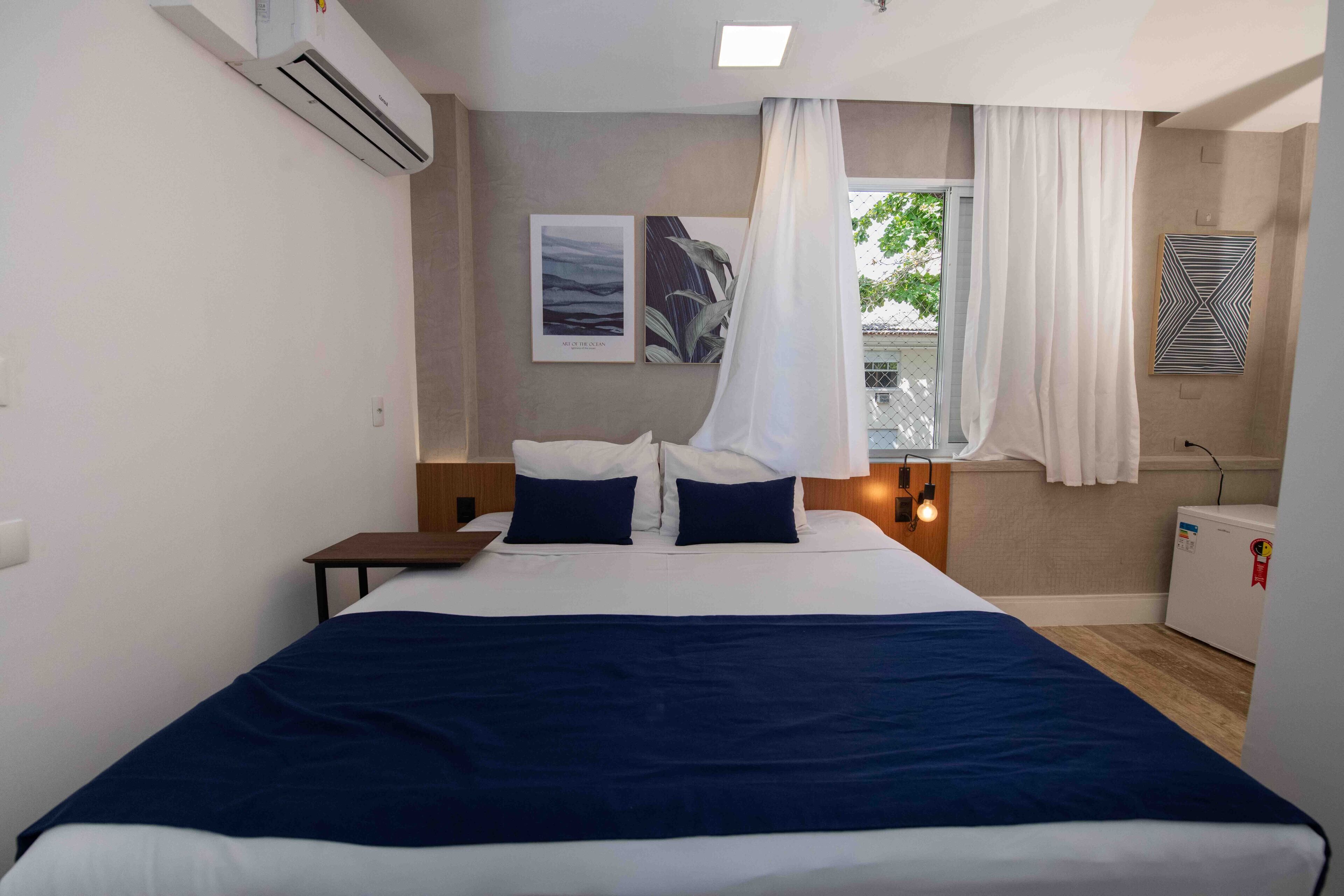 A hotel room with a bed, air conditioning unit, artwork, a window with curtains, and a bedside table.