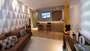 A modern living room with a brown sofa, two bar stools, a TV mounted on the wall, and a wooden counter. - Leblon Design Hotel (Rio de Janeiro)
