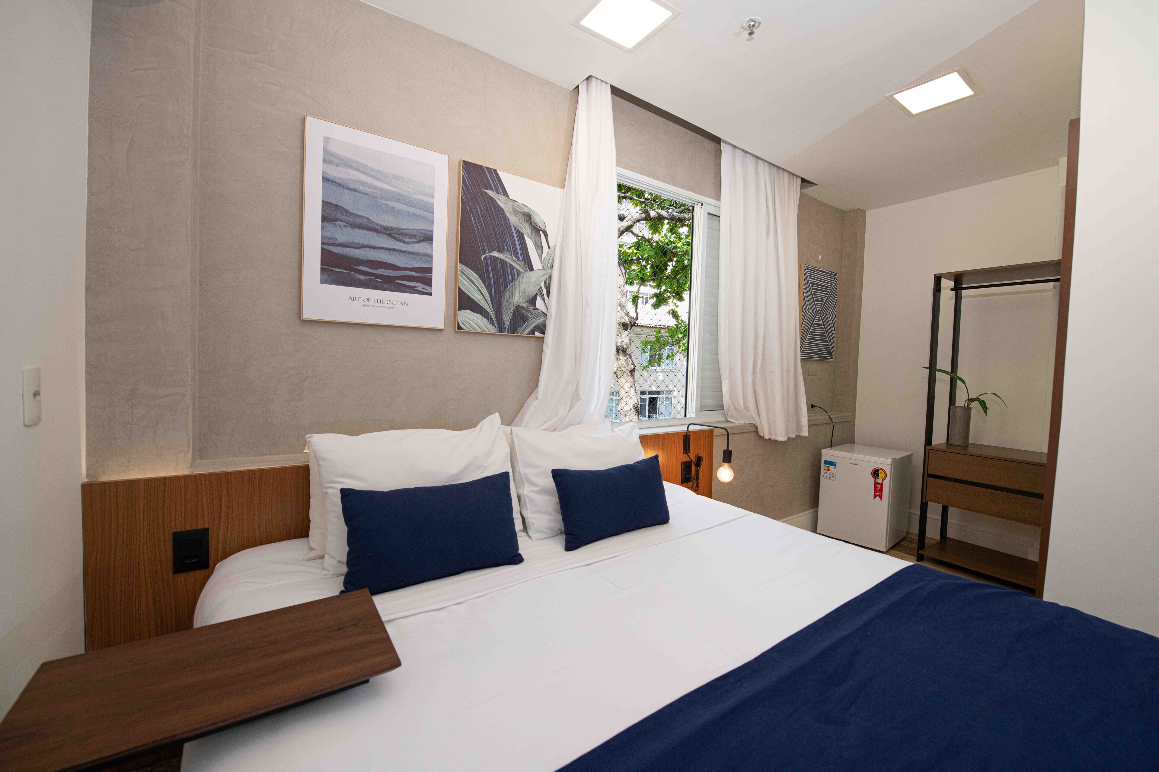 A hotel room with a bed, a nightstand, a small table, a mirror, a window with curtains, and artwork on the wall.