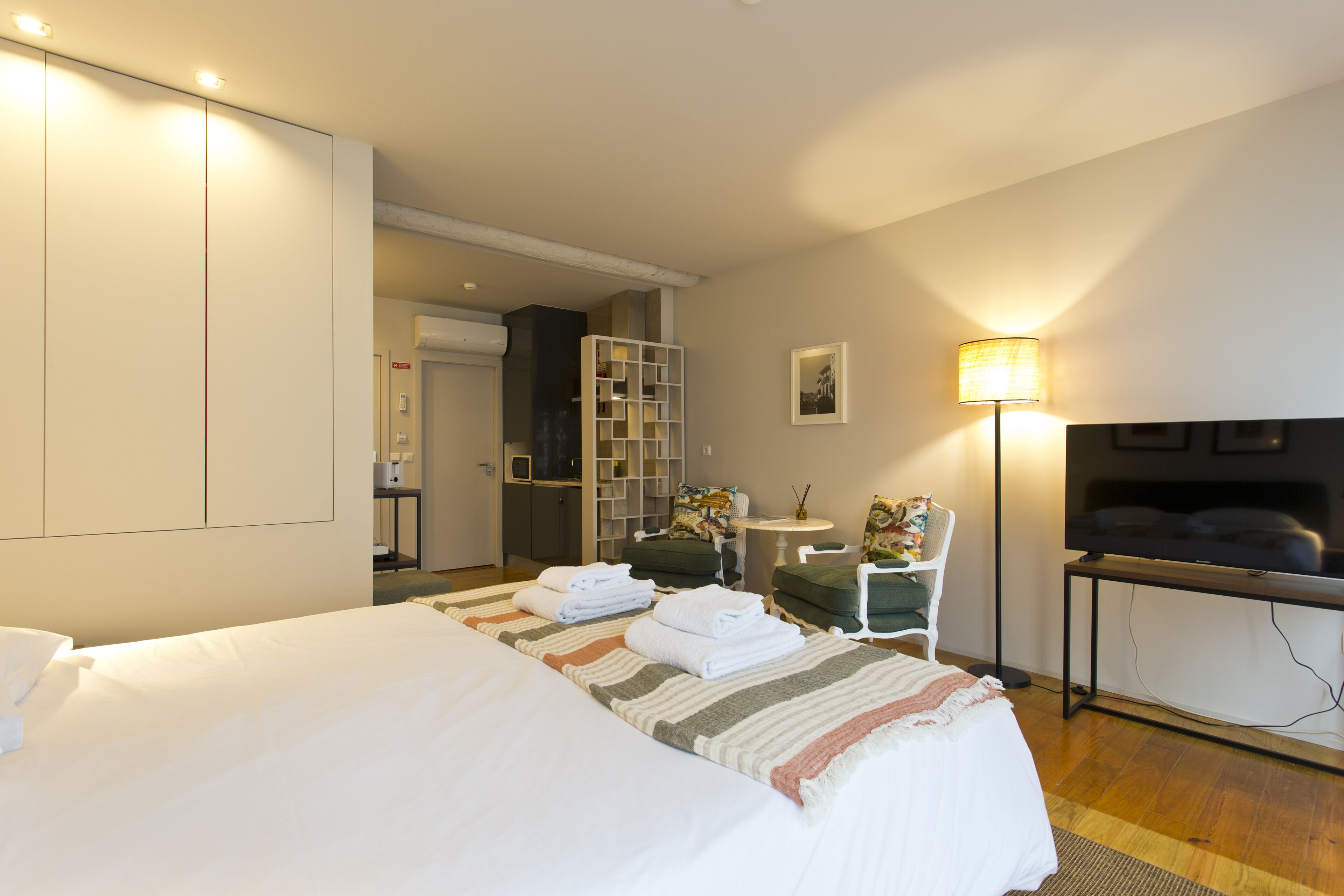 A hotel room with a large bed, a desk with a flat-screen TV, a chair, a small table, and a lamp.