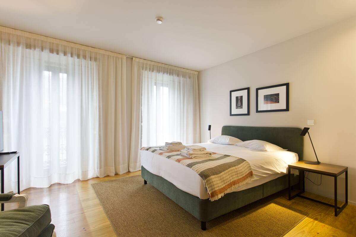 A neatly made bed with a green headboard, a bedside table with a lamp, and a large window with sheer curtains.