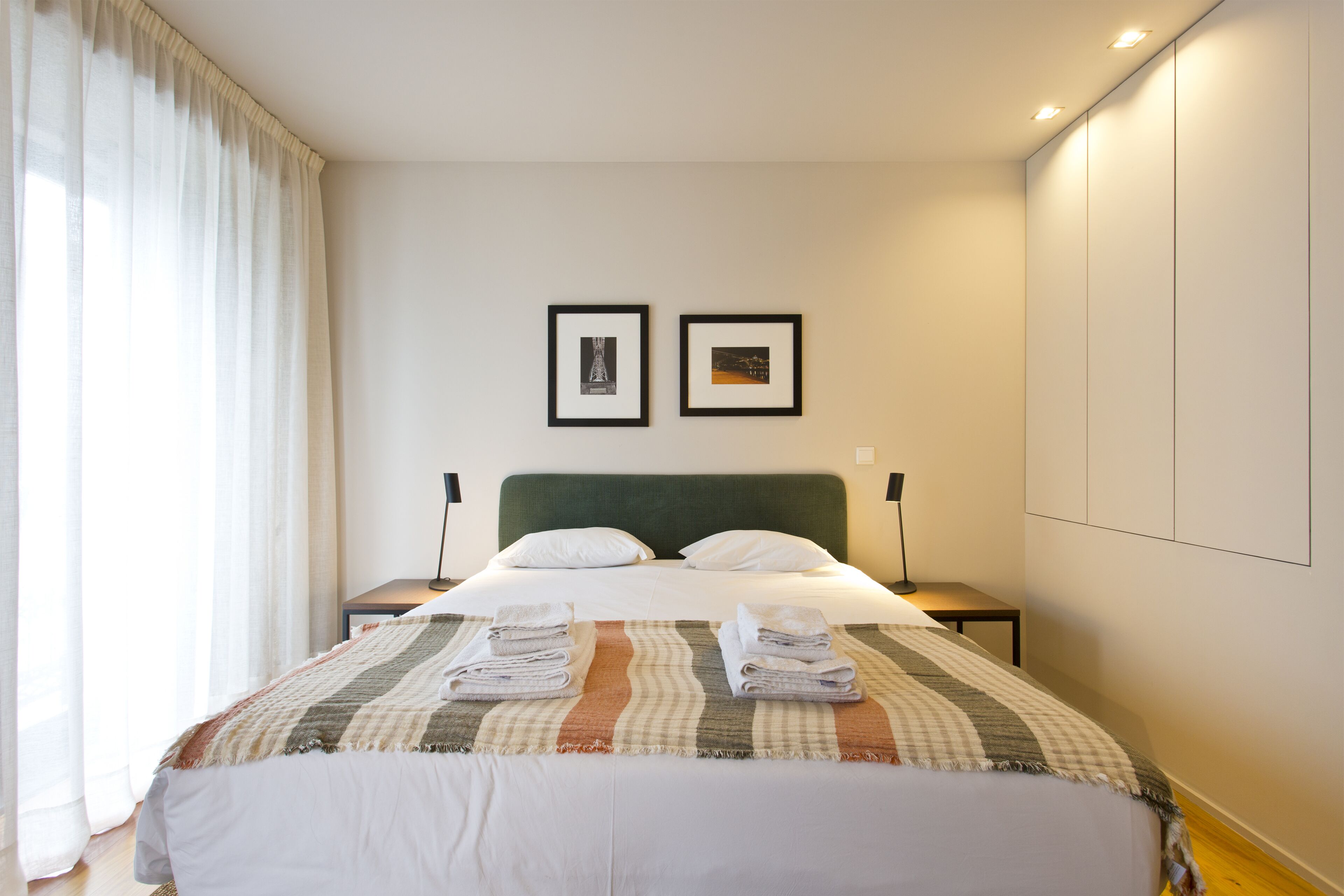 A neatly made bed with a striped blanket, two framed pictures on the wall, and a bedside table with a lamp.