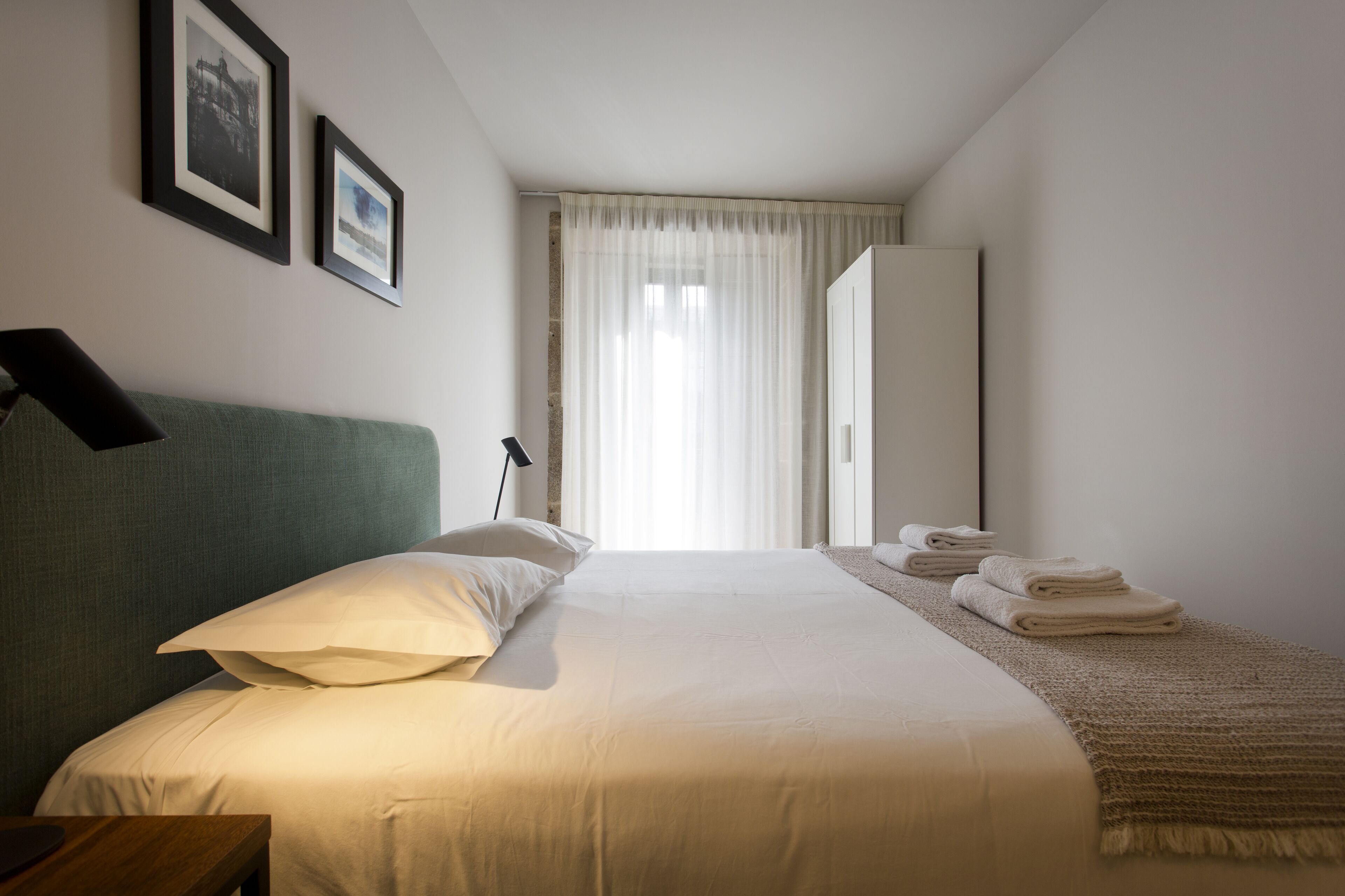 A neatly made bed with pillows, a bedside table, and a lamp. There are two framed pictures on the wall, a window with sheer curtains, and a folded towel on the floor.
