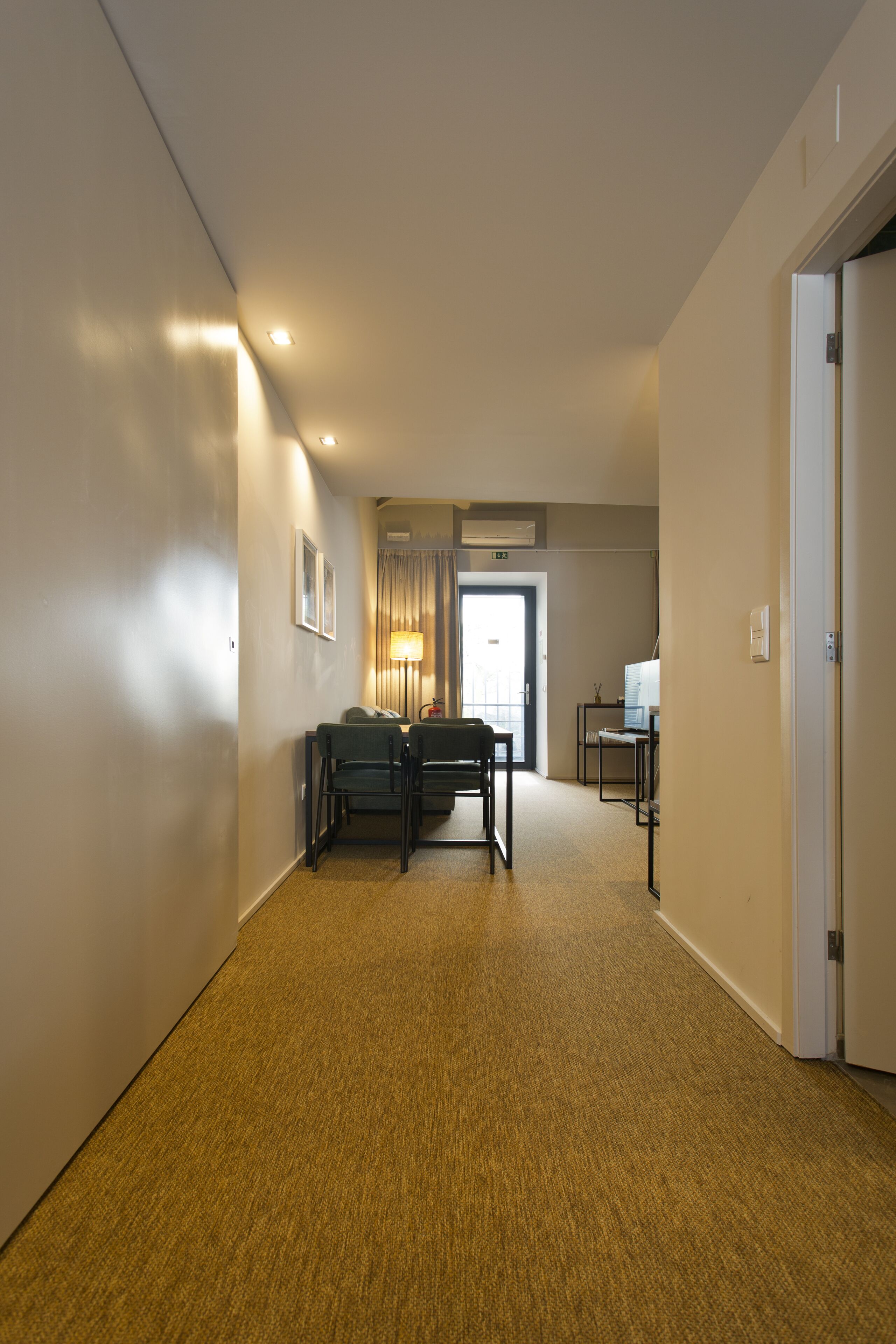 Un couloir doté d’un revêtement de sol en moquette, un bureau avec des chaises, une lampe et une porte donnant sur une autre pièce.
